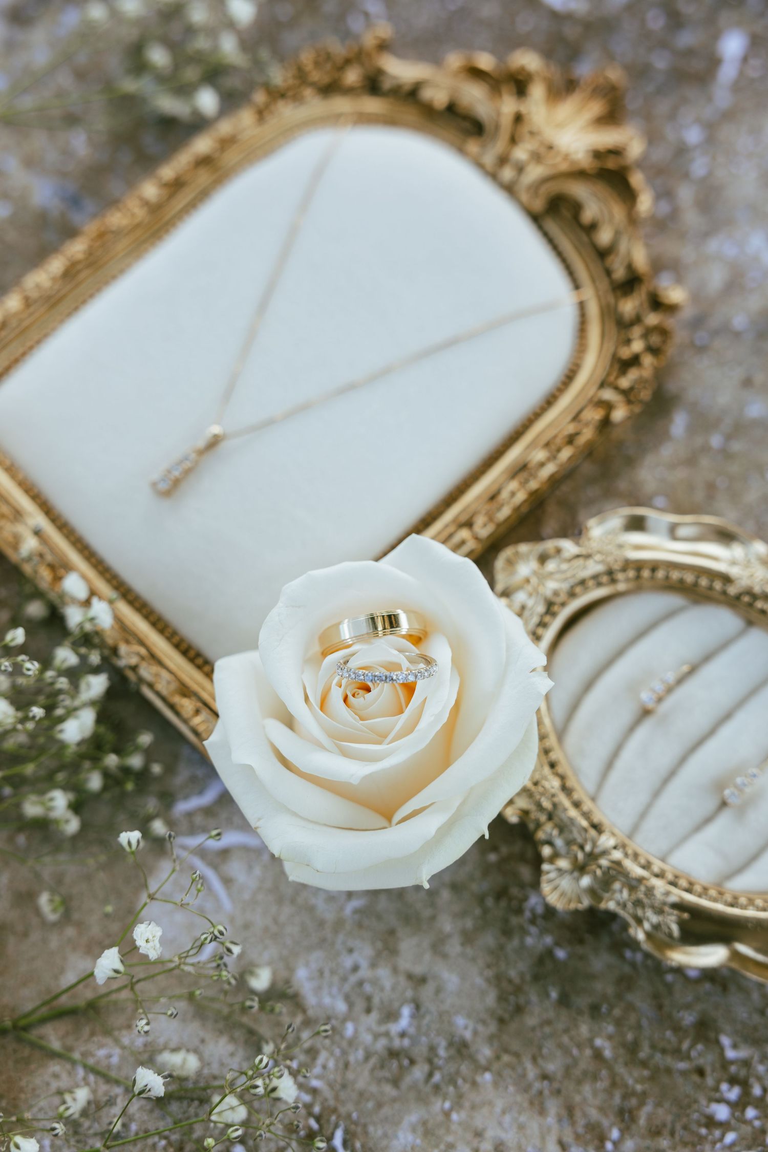 Gold wedding rings rest on a white rose beside ornate golden frames displaying a diamond necklace and earrings with baby's breath flowers.