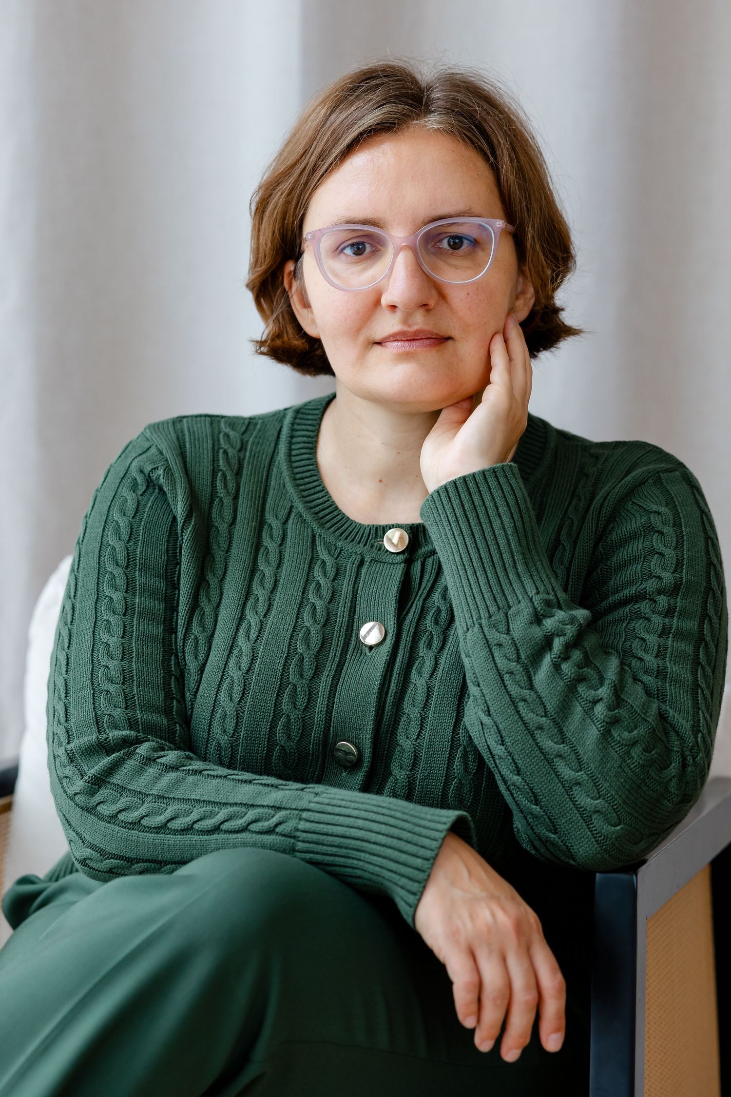 Woman in green cable-knit cardigan with glasses sits thoughtfully, resting chin on hand against light curtain backdrop.