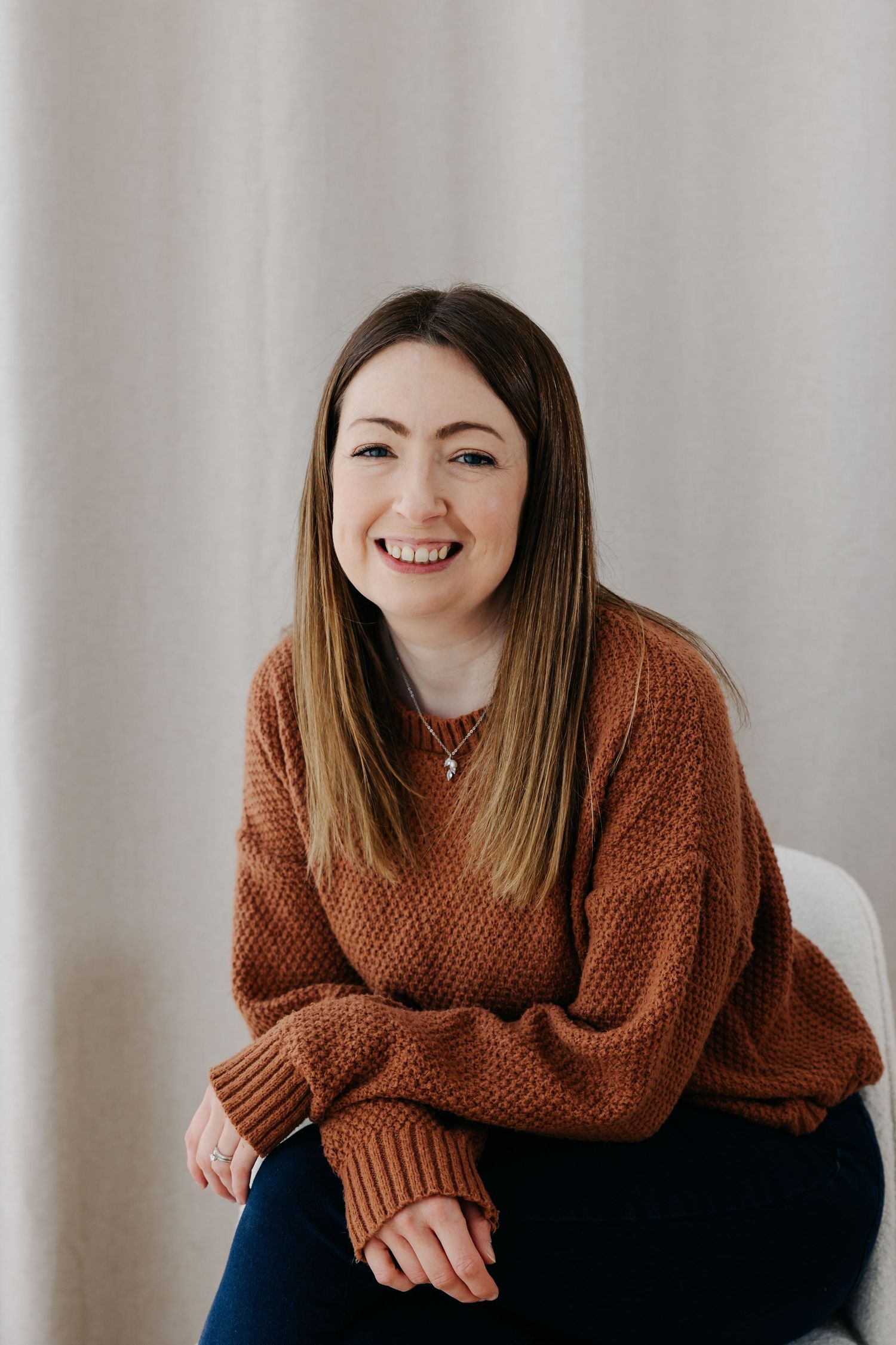 Smiling young woman with long brown hair wearing a rust-colored knit sweater, seated casually against a light curtain backdrop.