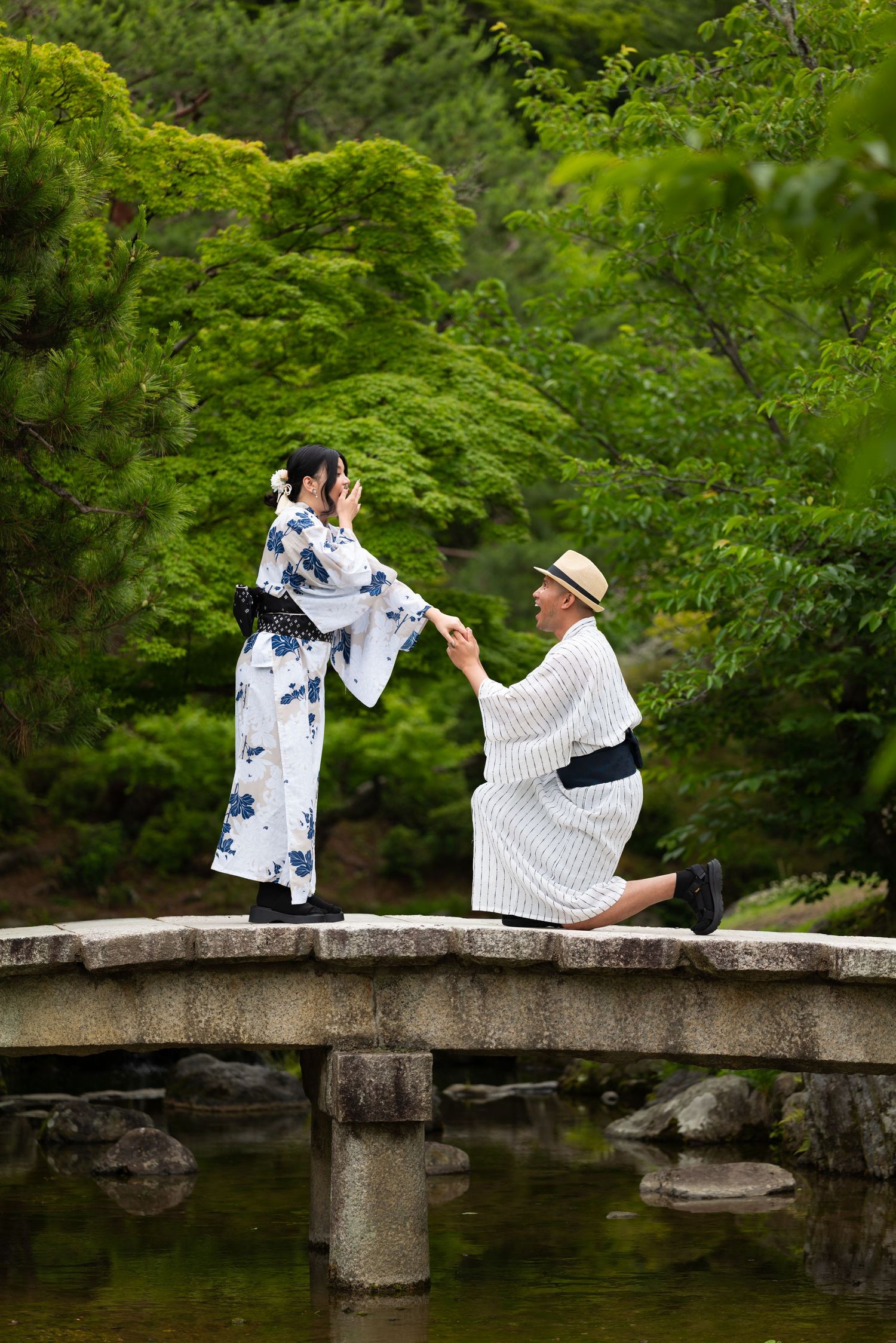 Couple posing with arms outstretched on a stone bridge over a pond in a lush Japanese garden.