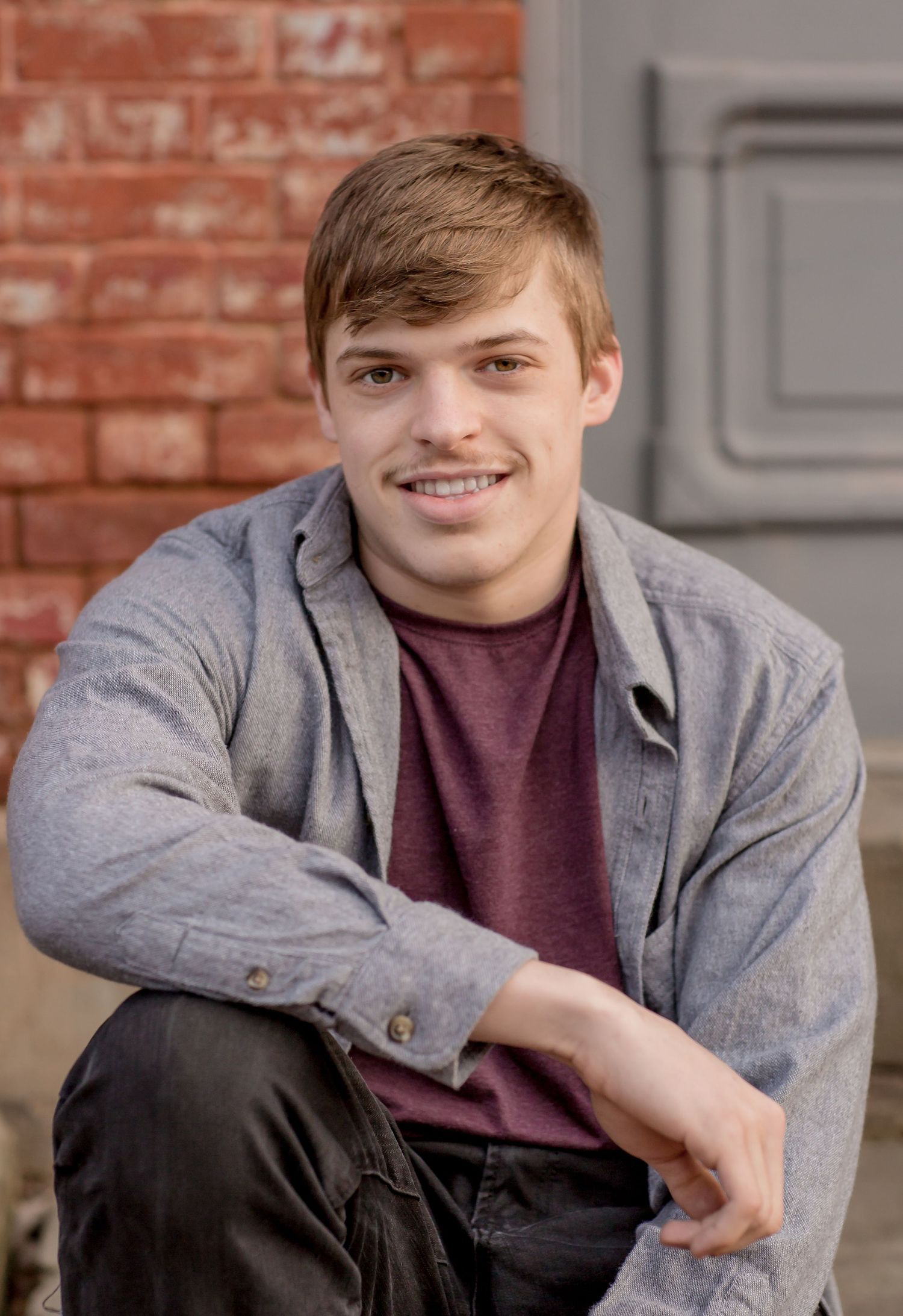 Smiling young man in a gray shirt and maroon tee sitting casually in front of a red brick wall.