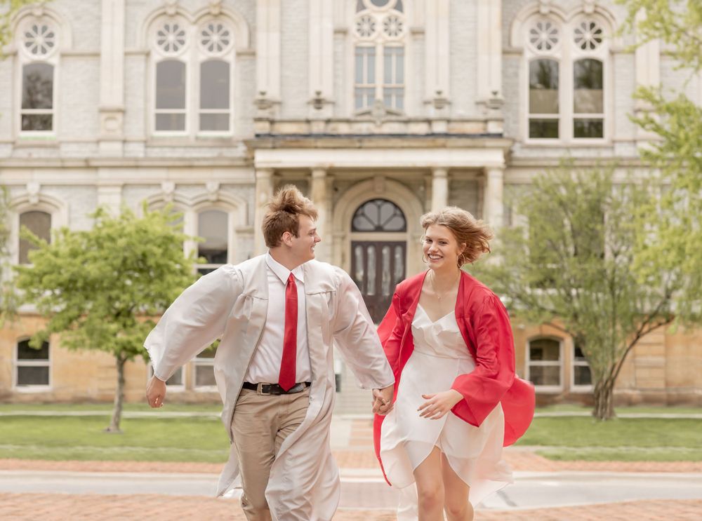 Two graduates in red and white regalia run playfully in front of historic building.