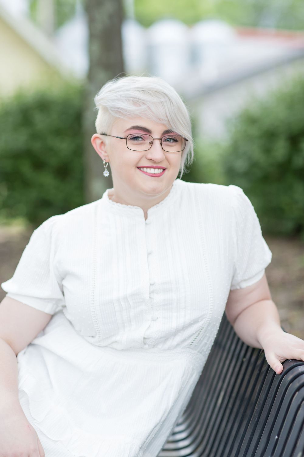 Young woman with short platinum hair and glasses smiles while seated on outdoor bench.