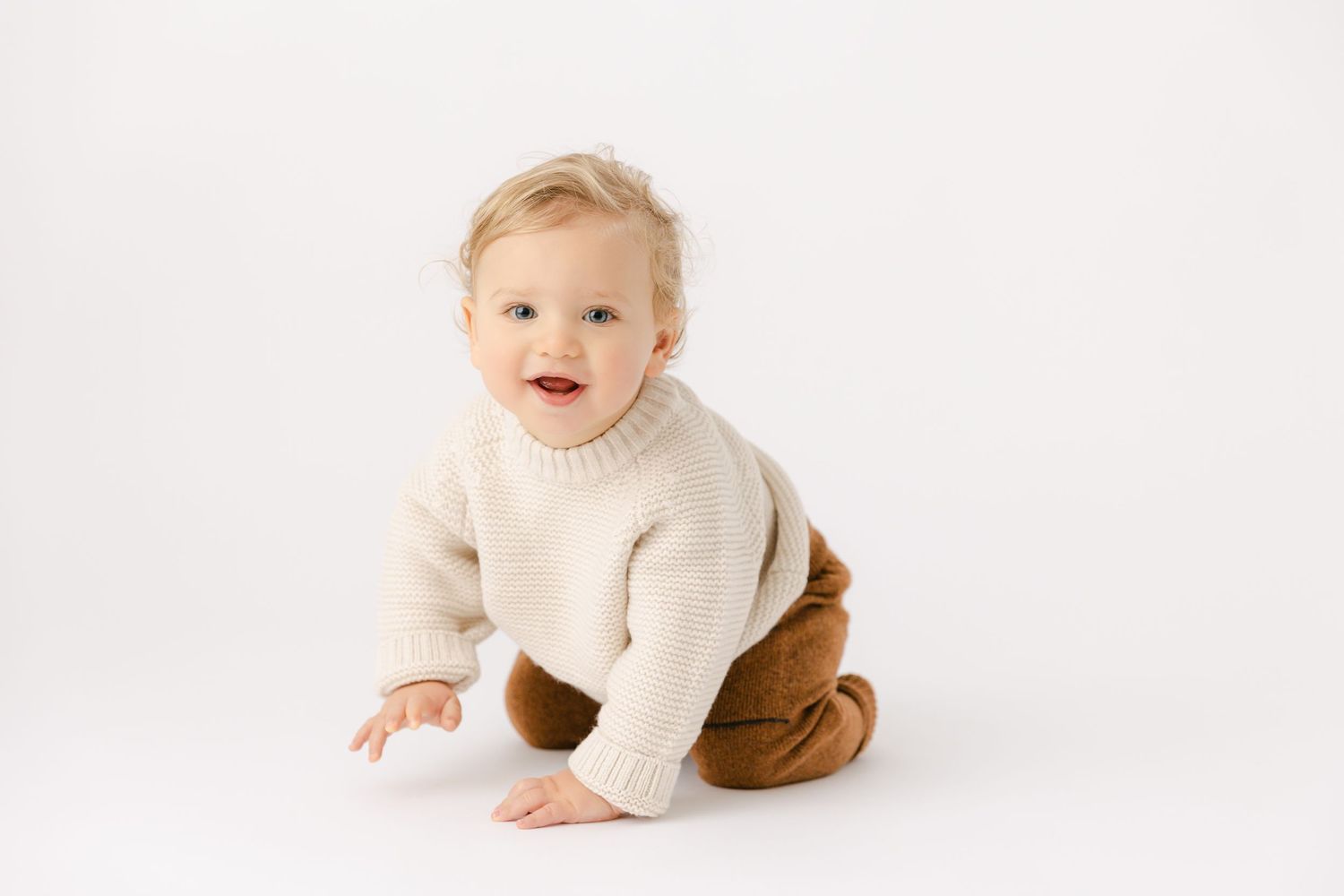Blonde haired baby wearing a cream knit sweater and brown pants smiles and crawls toward the camera.