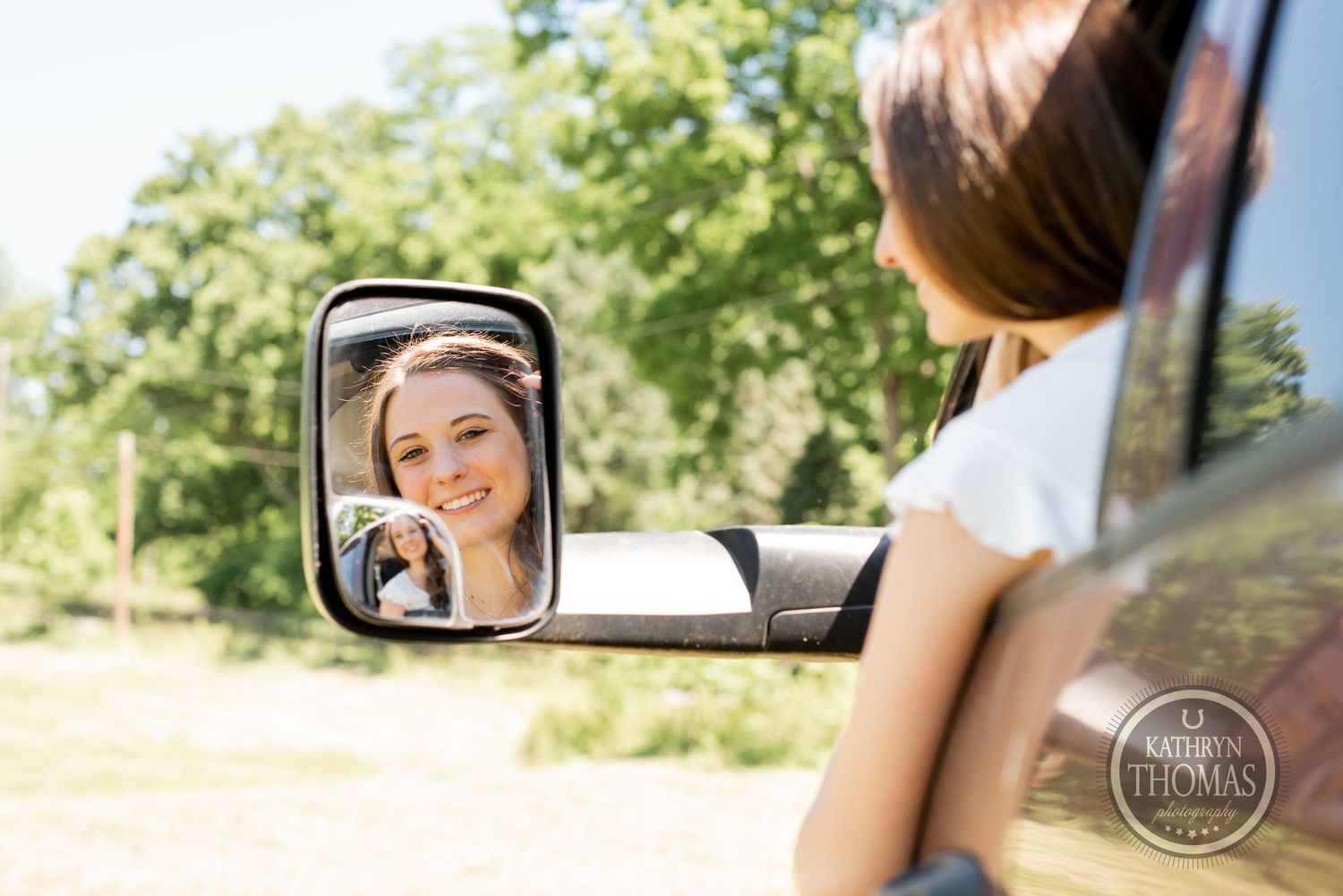 A person's reflection in a car side mirror on a sunny summer day with trees in the background.