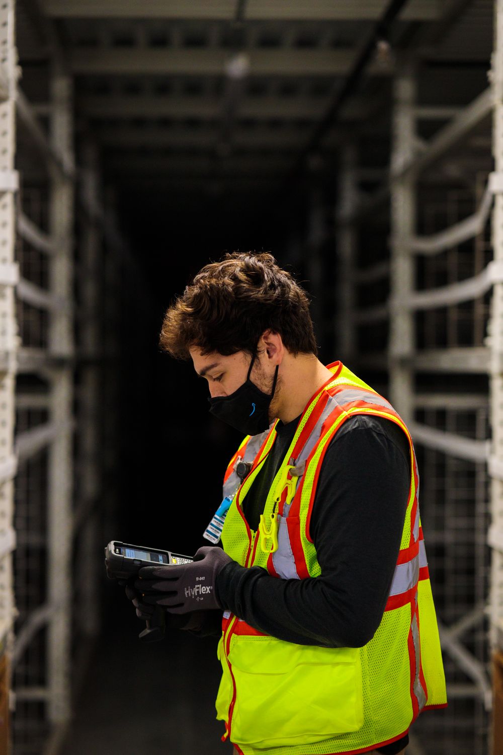 A man in a mask and vest stands in an Amazon warehouse, surrounded by shelves and boxes during their new warehouse grand opening