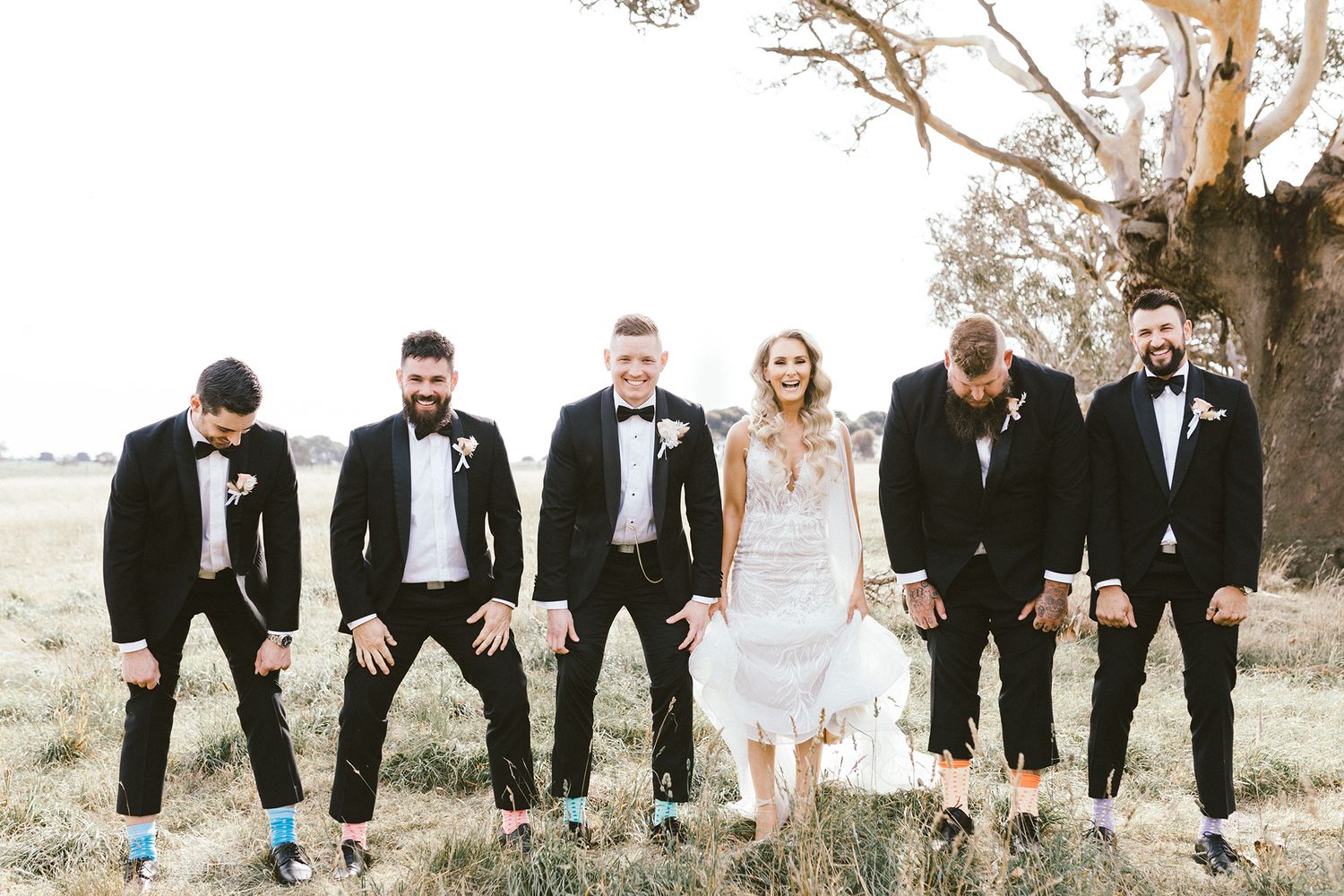 Playful wedding party shows off colorful socks while standing together in a field.