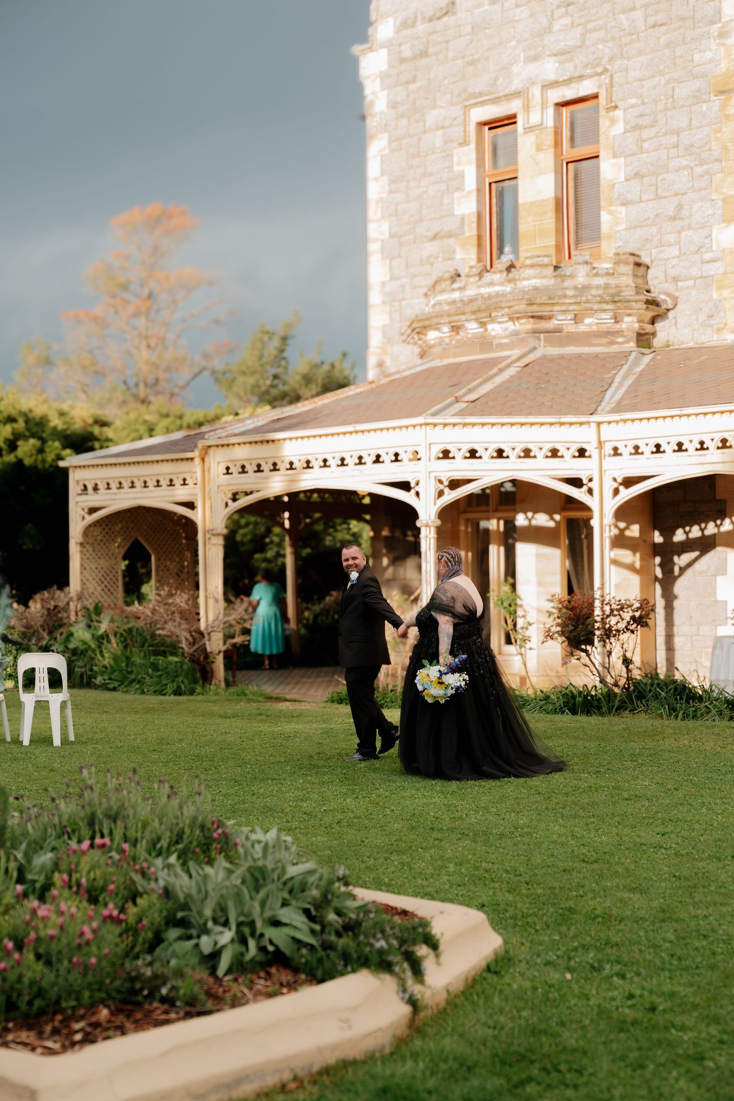 Bride in black wedding gown holds hands with groom outside historic stone mansion with ornate veranda under dramatic sky.