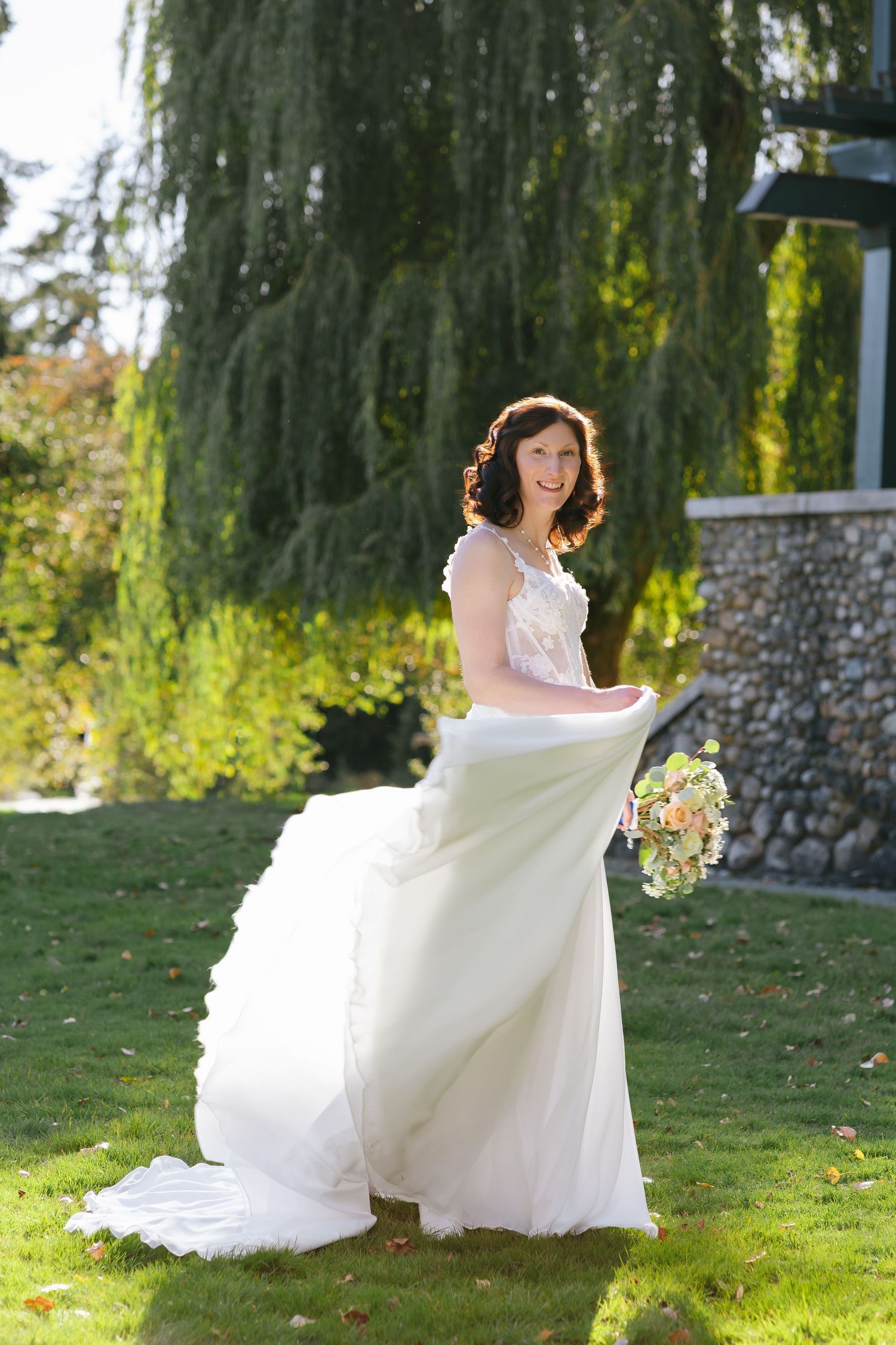 Smiling bride in a flowing white wedding gown holding a pastel bouquet on a sunlit lawn beside a weeping willow and stone wall.