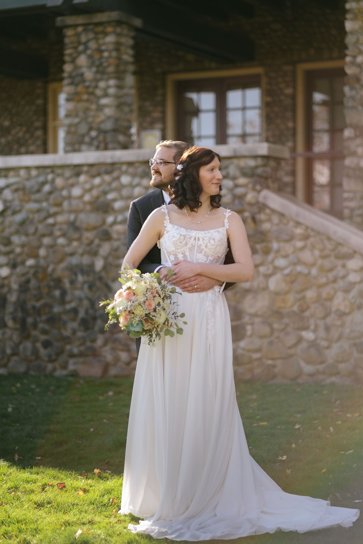 Michael embracing Melissa from behind as she holds her bouquet in front of a stone building at Deer Lake Park.