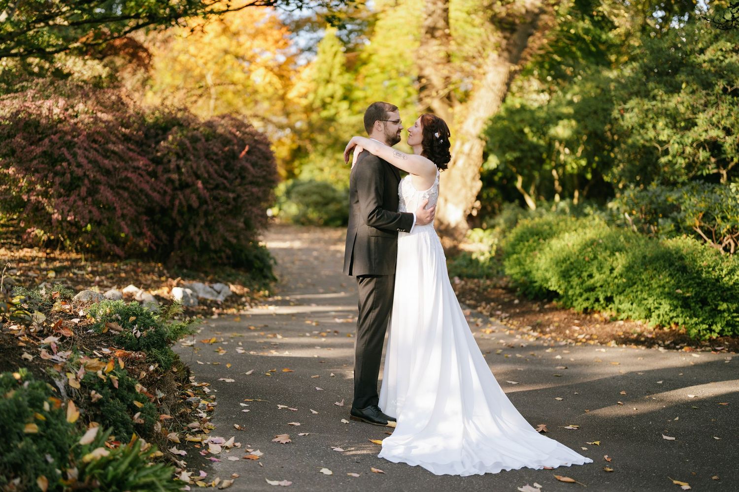 Michael and Melissa kissing on a tree-lined path at Deer Lake Park, surrounded by autumn foliage.