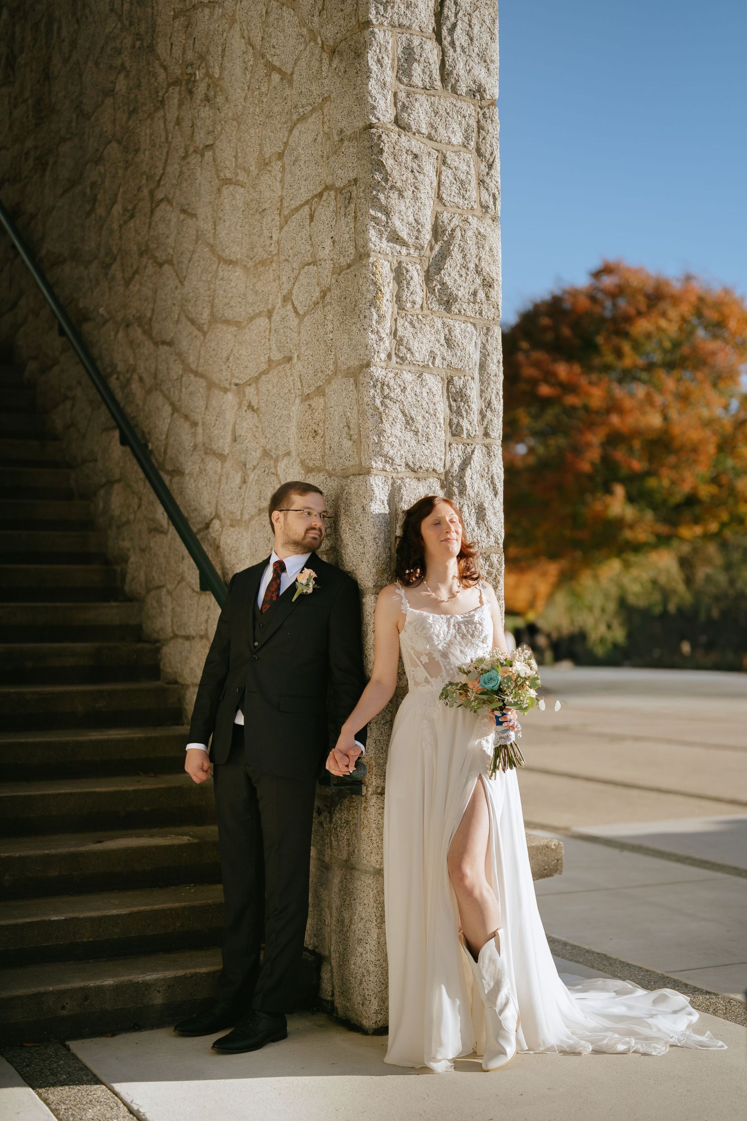 Bride in a white gown and cowboy boots holds hands with a groom in a dark suit beside a stone pillar and steps on an autumn day.