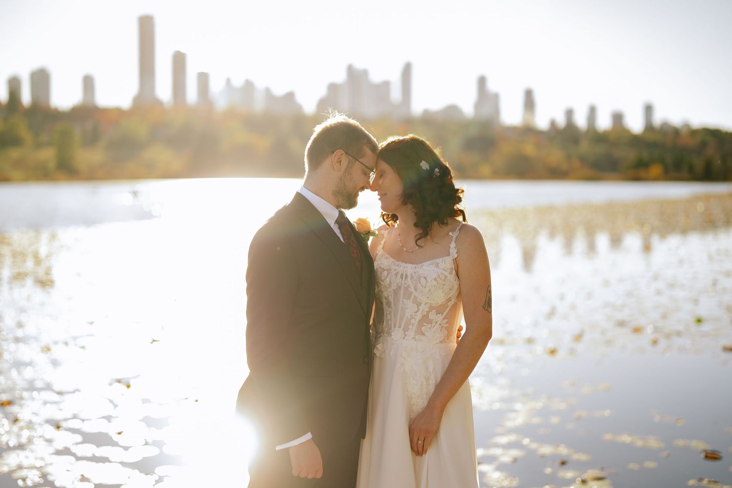 Michael and Melissa touching foreheads beside the sunlit lake at Deer Lake Park, with a hazy city skyline behind them.