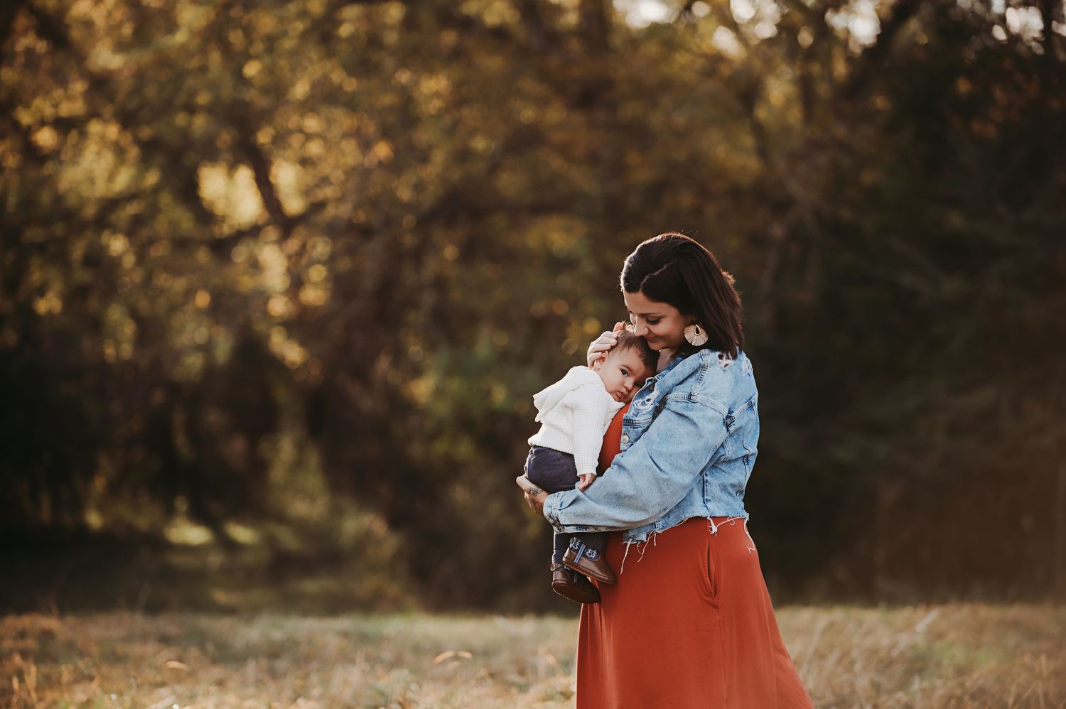 A woman in a denim jacket and rust orange skirt tenderly hugs her baby while standing in a sunlit autumn field with golden trees behind her.