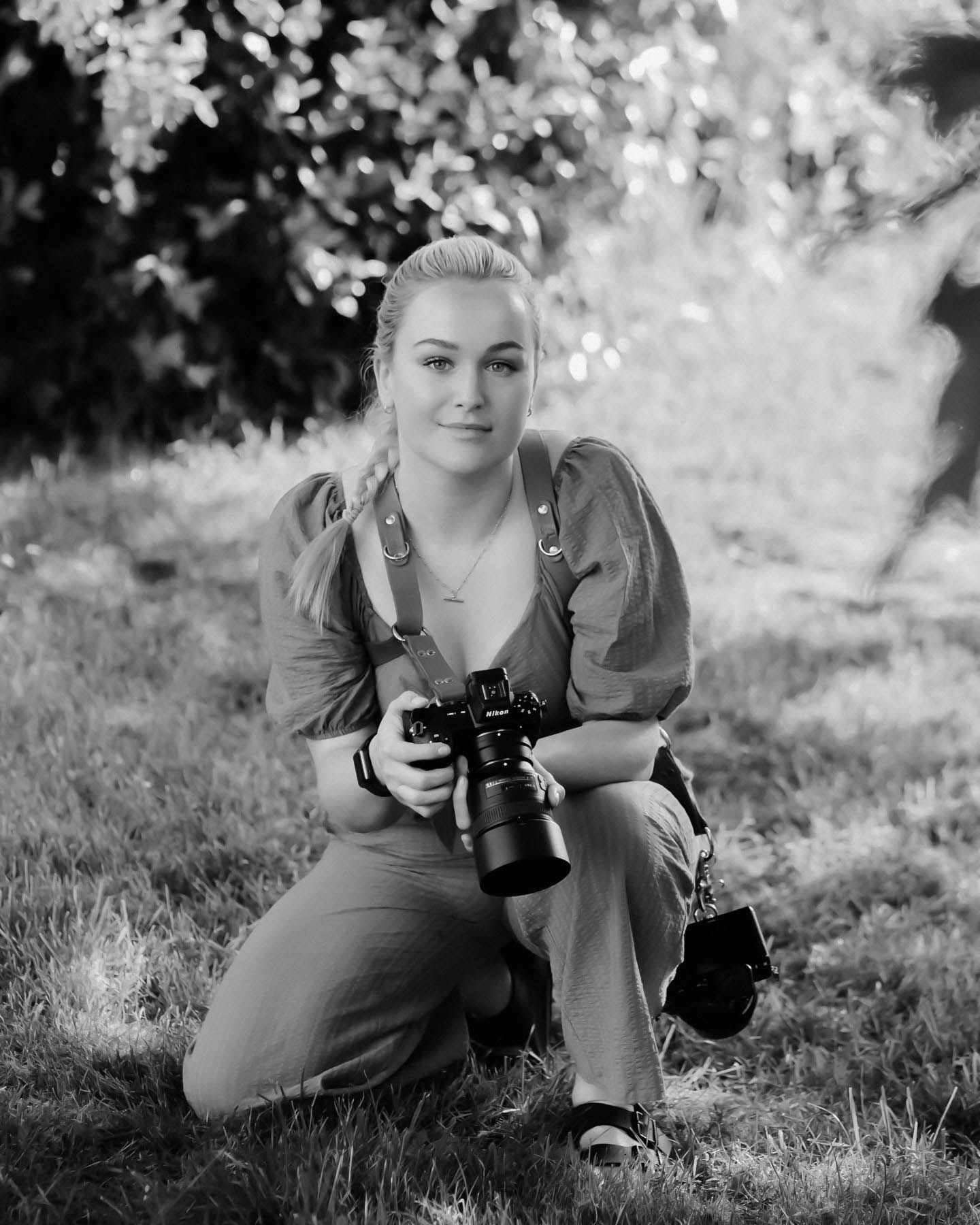 A photographer in casual attire kneels with camera equipment in an outdoor setting.