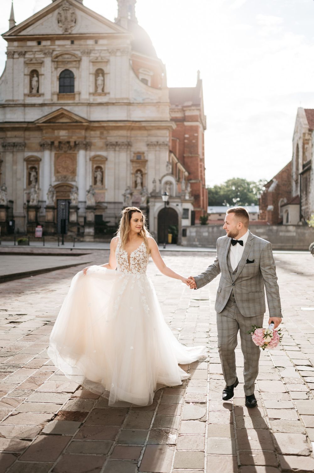Elegant couple walking hand in hand in front of historic church with sunlight peeking through.