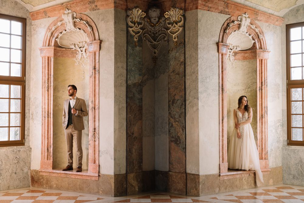 Couple in wedding attire posed in symmetrical baroque palace alcoves with ornate architectural details.
