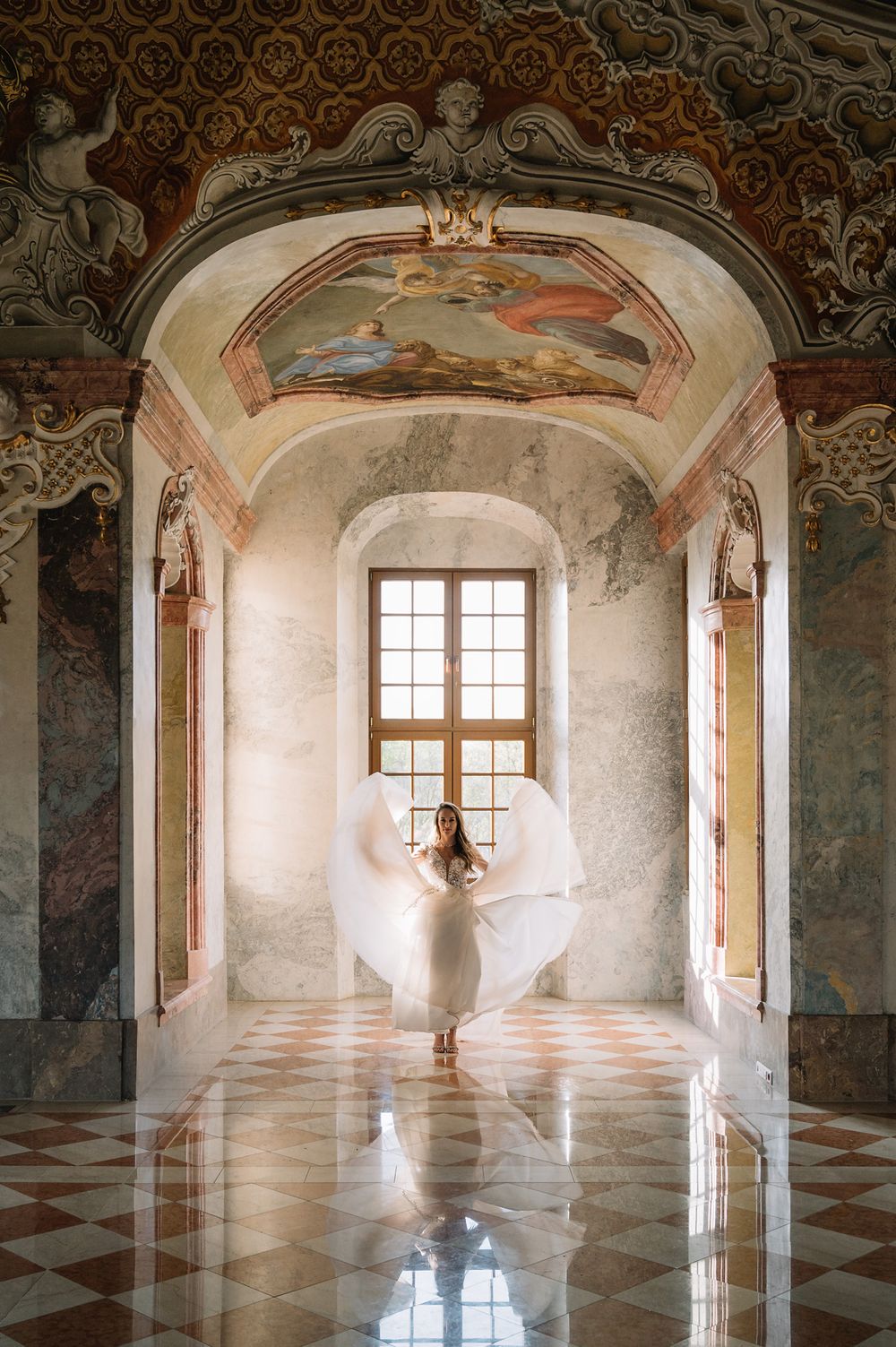 Figure in flowing white dress posed beneath ornate frescoed ceiling and arched windows in baroque palace.