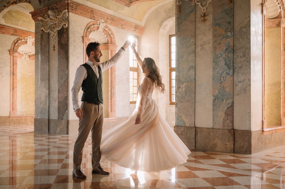 Couple dancing in elegant baroque palace hall with marble floors and decorated columns.