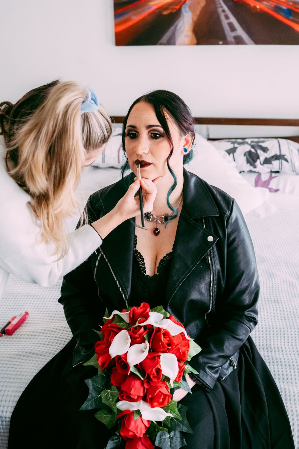 Makeup artist applies finishing touches to gothic bride holding red and white bouquet.
