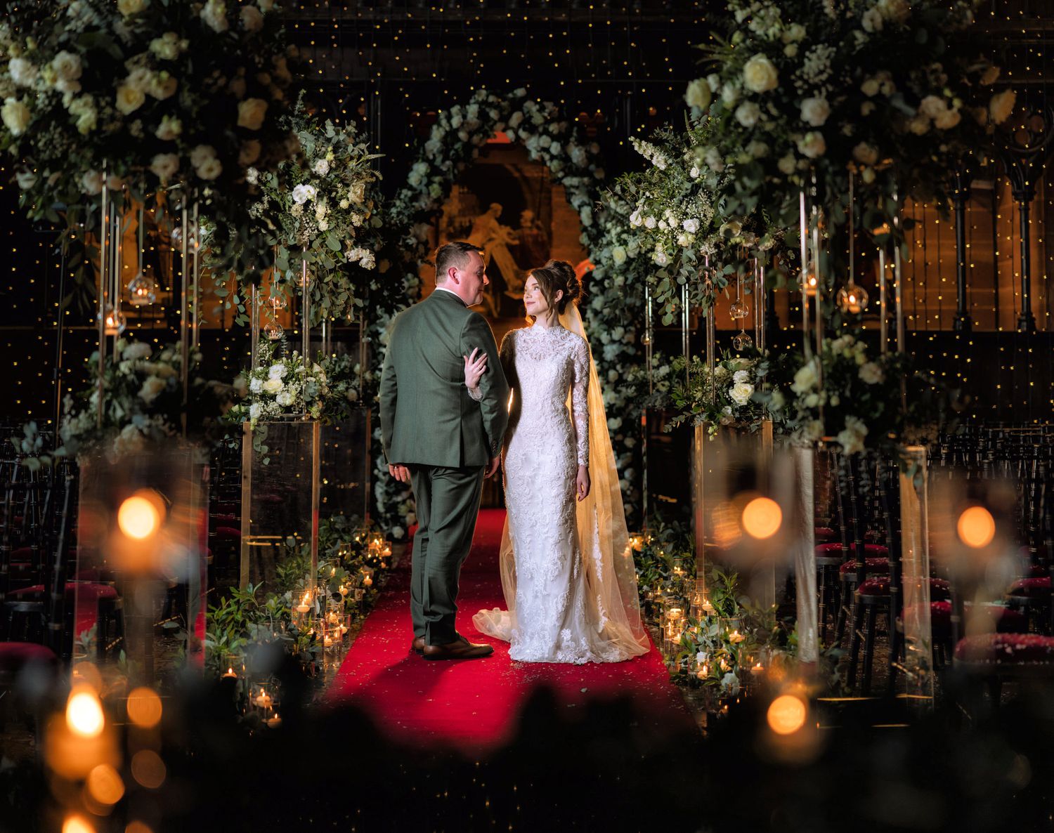 Bride in a lace gown and groom embrace on a red aisle runner beneath floral arches and glowing candlelight at Peckforton Castle wedding.