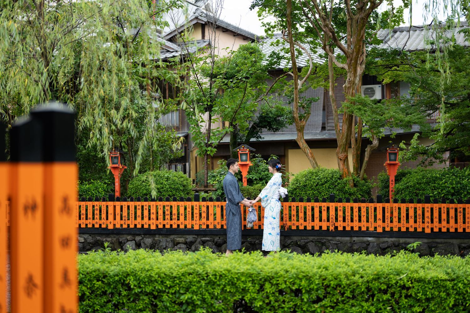 Couple in traditional kimono stand by an orange wooden fence among lush green trees and hedges in a peaceful Kyoto-style garden.