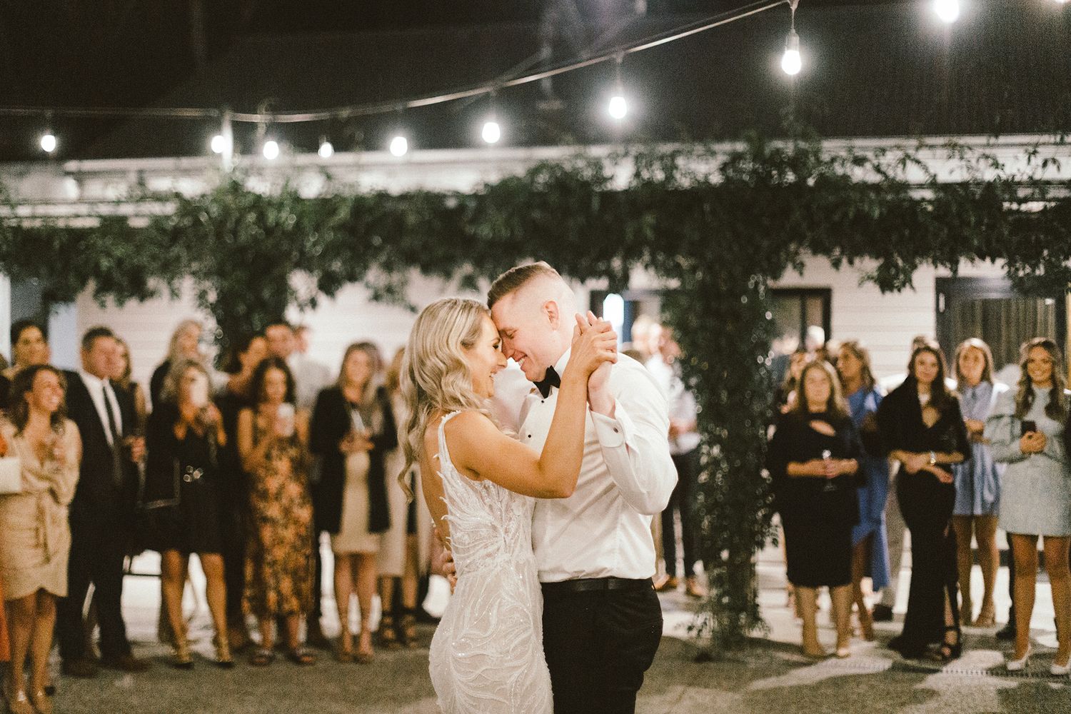 Evening wedding celebration under string lights with guests dancing outdoors.