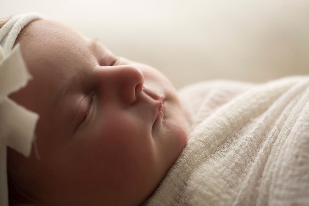 Newborn baby girl in white wrap and white headband backlit showing her facial details.
