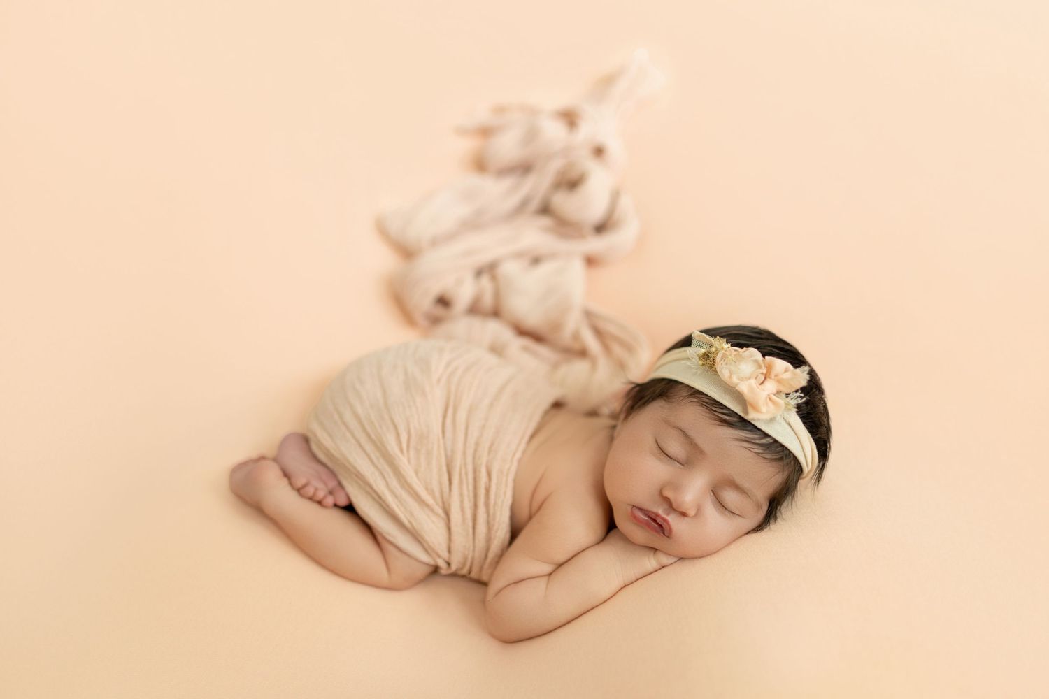 Sleeping newborn baby wrapped in cream fabric with floral headband on beige background.
