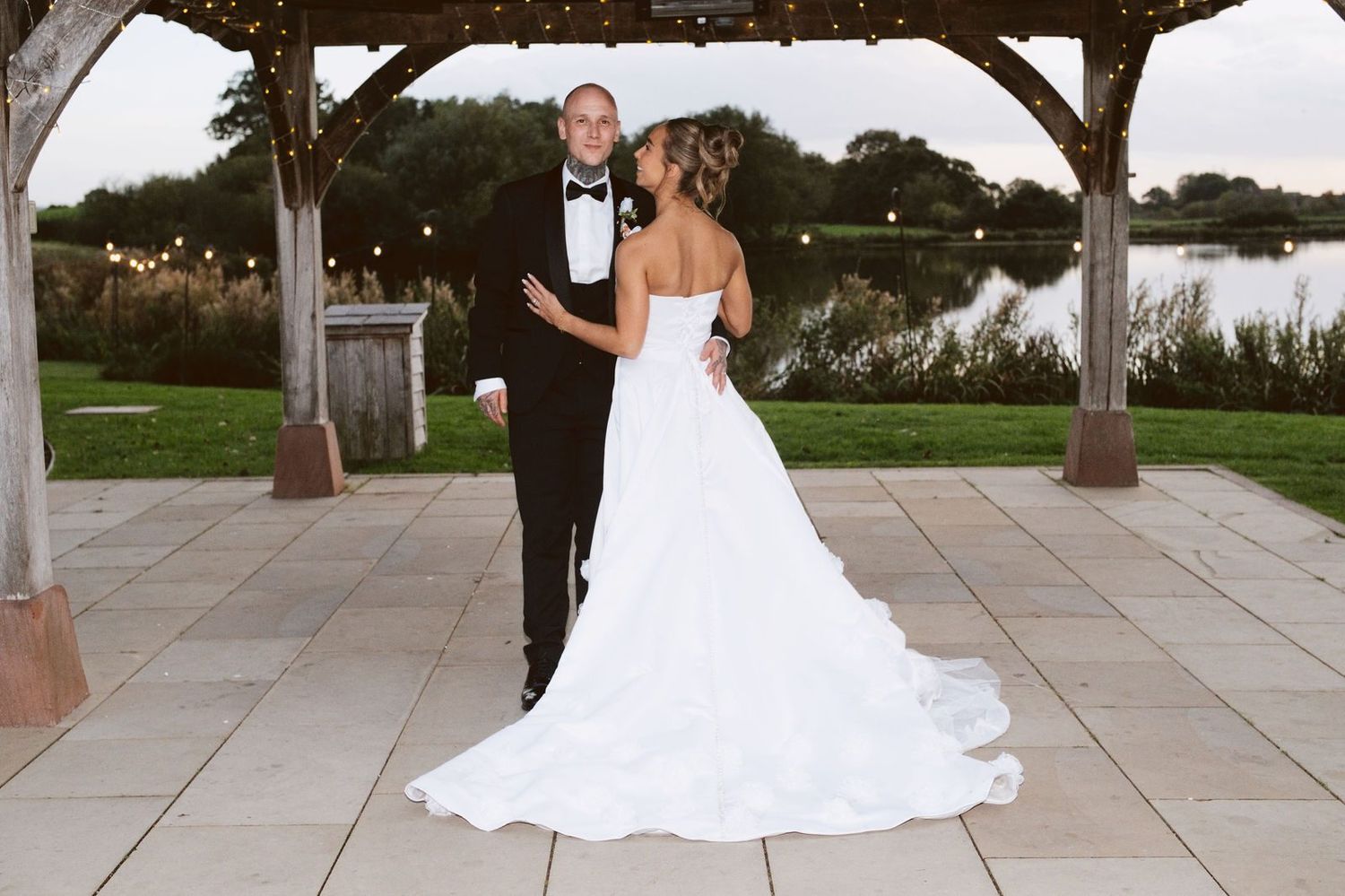 Bride and groom under the lakeside gazebo with festoon lights and the lake behind, Sandhole Oak Barn wedding