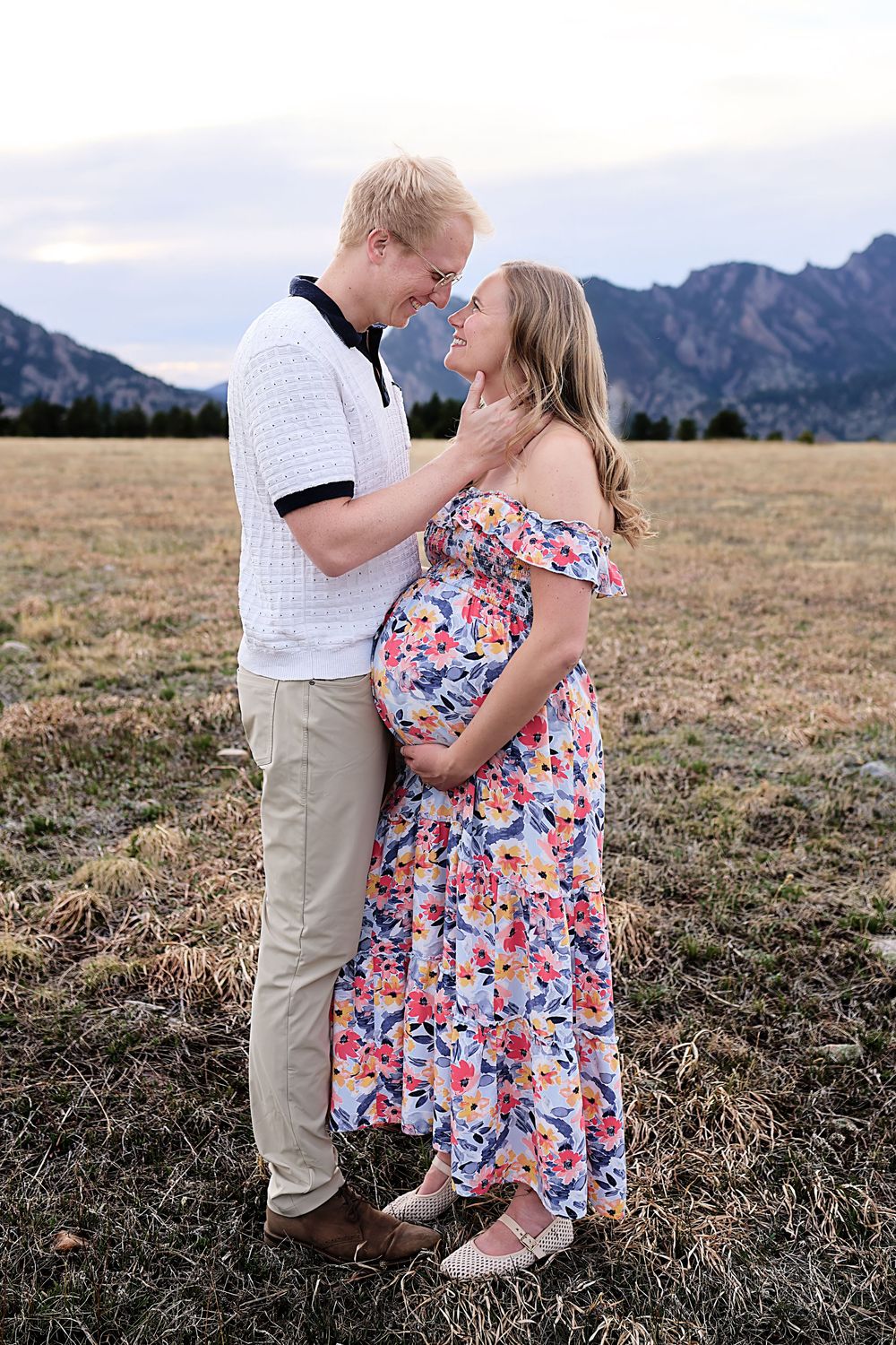 Smiling couple embracing in a grassy field with mountains behind, the pregnant woman in a floral dress showing her baby bump at sunset.
