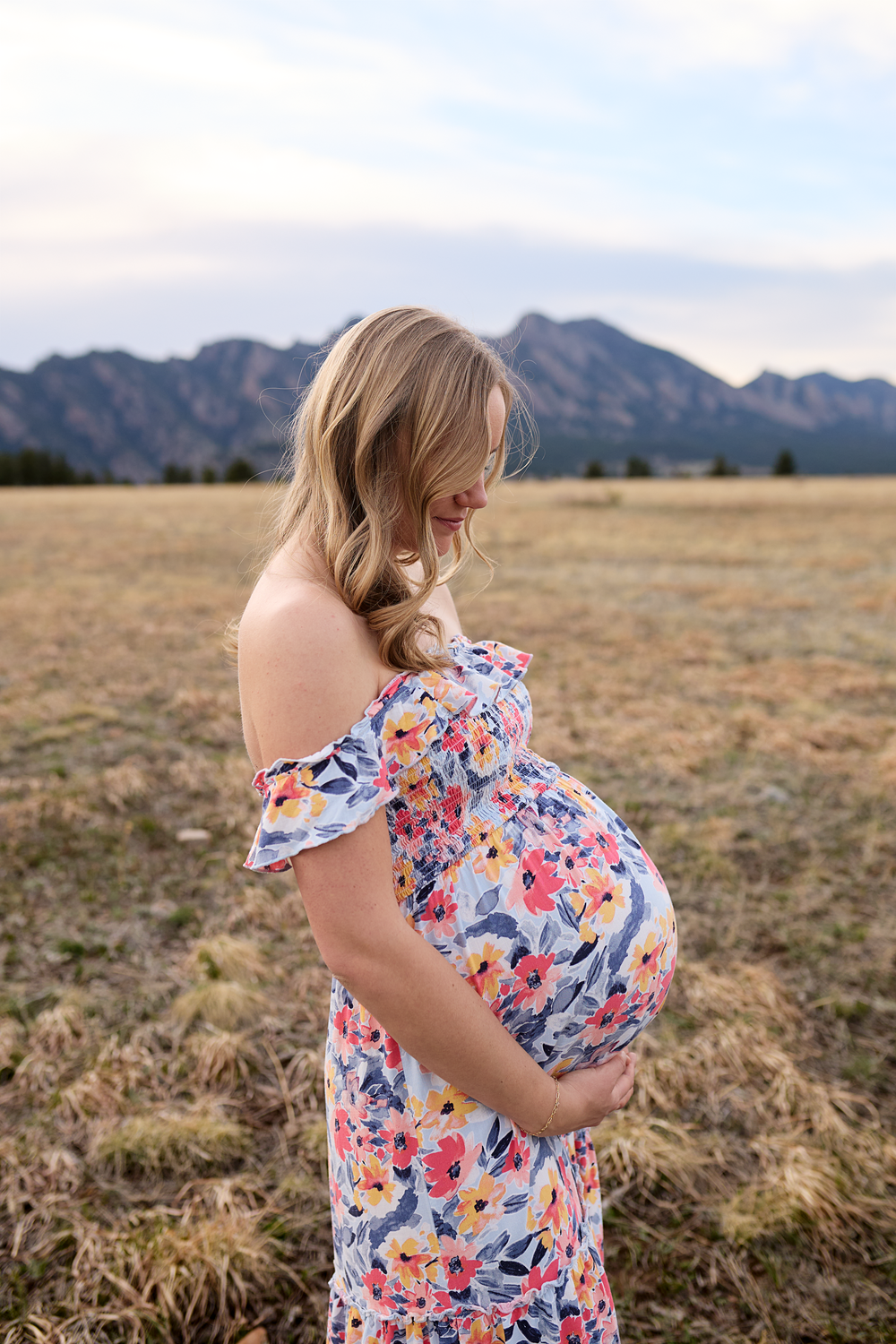 Pregnant woman in a floral off-the-shoulder dress cradles her baby bump in a grassy field with mountains in the background.