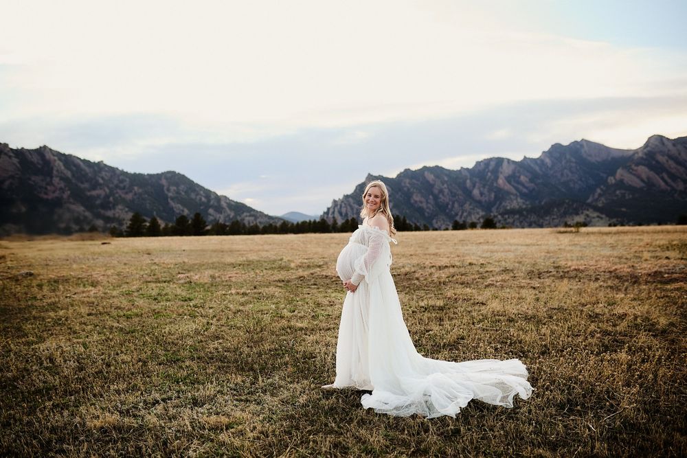 Pregnant woman in a flowing white gown with a long train stands in a golden grassy field with mountains behind her at sunset.