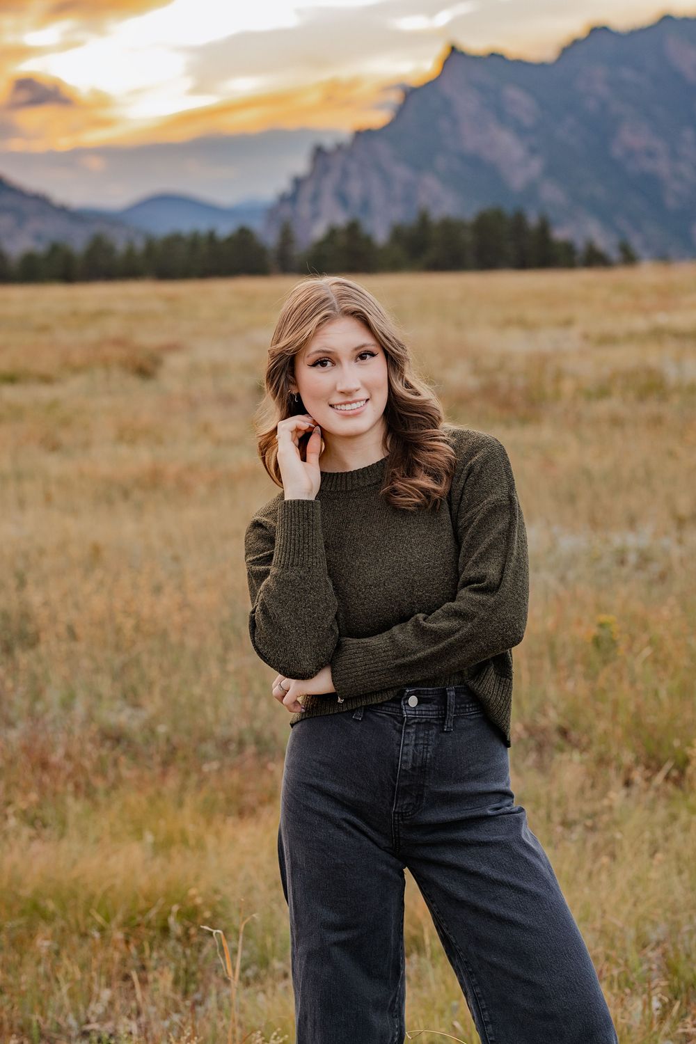 Smiling young woman in an olive green sweater and dark jeans poses in a golden meadow with mountains and a sunset sky behind her.