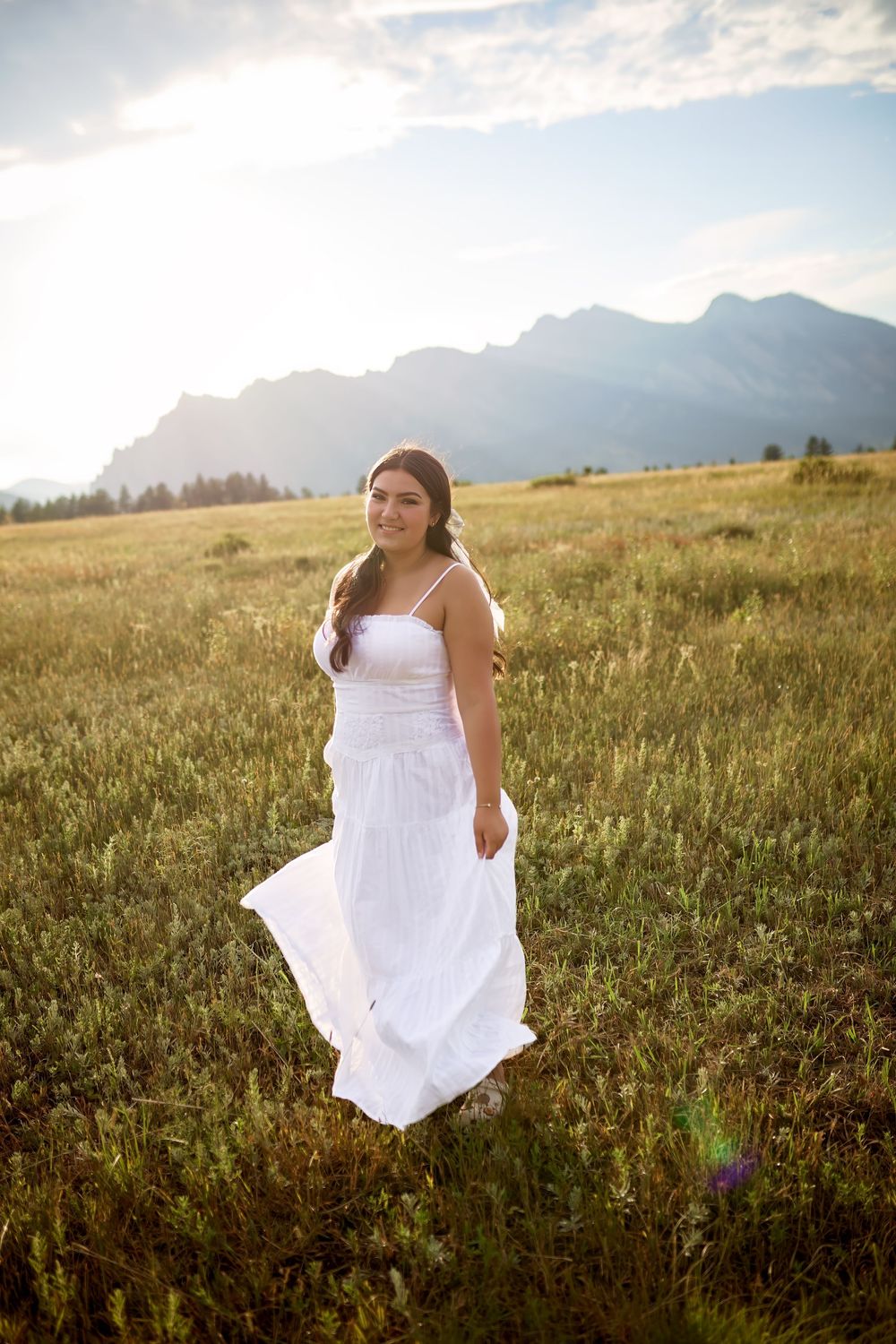 Smiling young woman in a flowing white dress stands in a golden grassy meadow with mountains and a sunlit sky behind her.