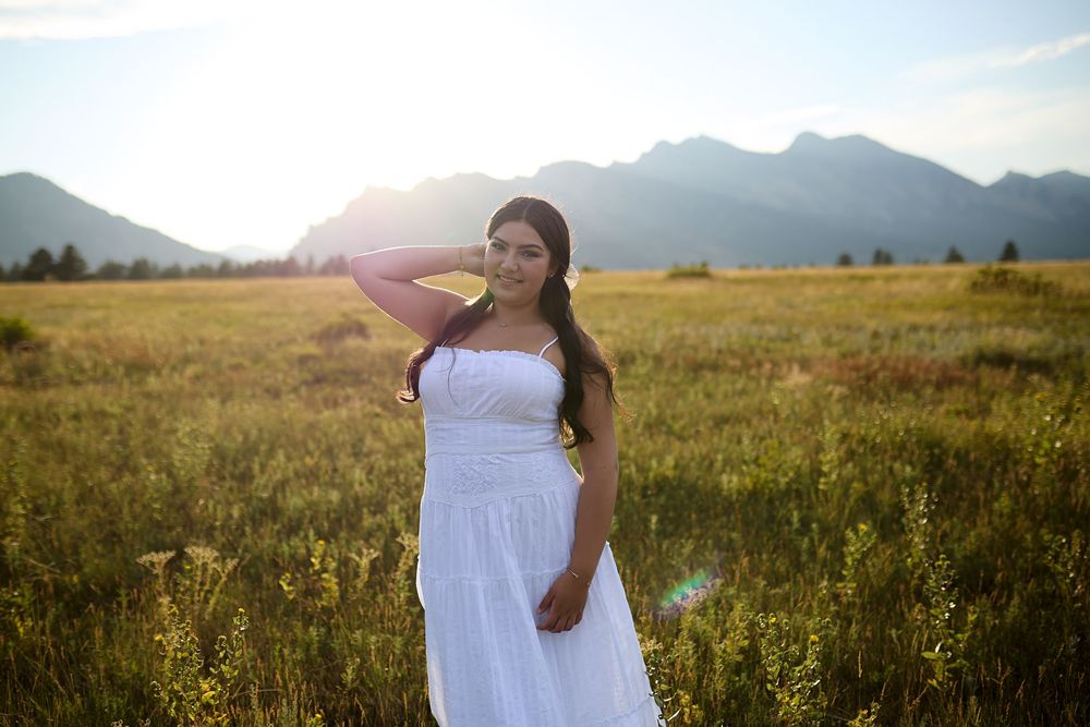 Woman in a white dress poses with one hand behind her head in a golden grassy meadow with mountains in the background at sunset.