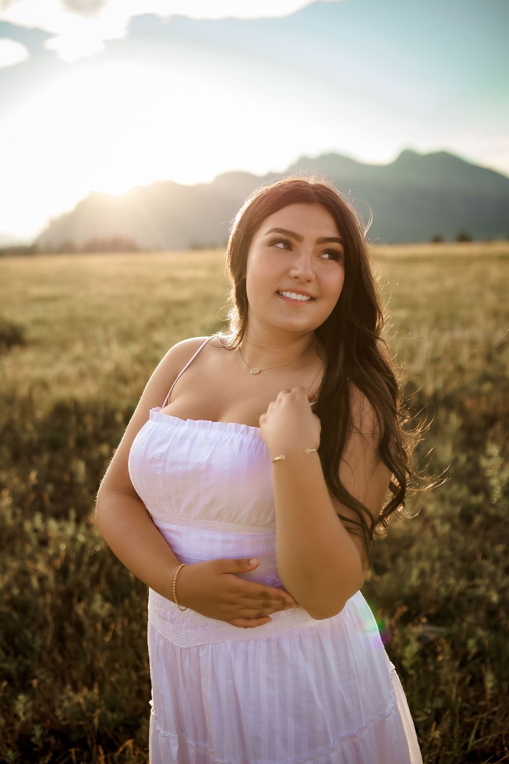 Smiling woman in a white sundress with long wavy hair stands in a golden grassy field at sunset with mountains in the background.