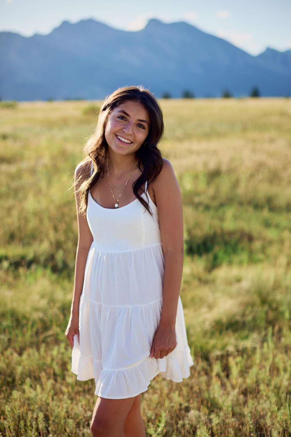 Smiling young woman in a white sundress stands in a golden grassy field with mountains in the background at sunset.