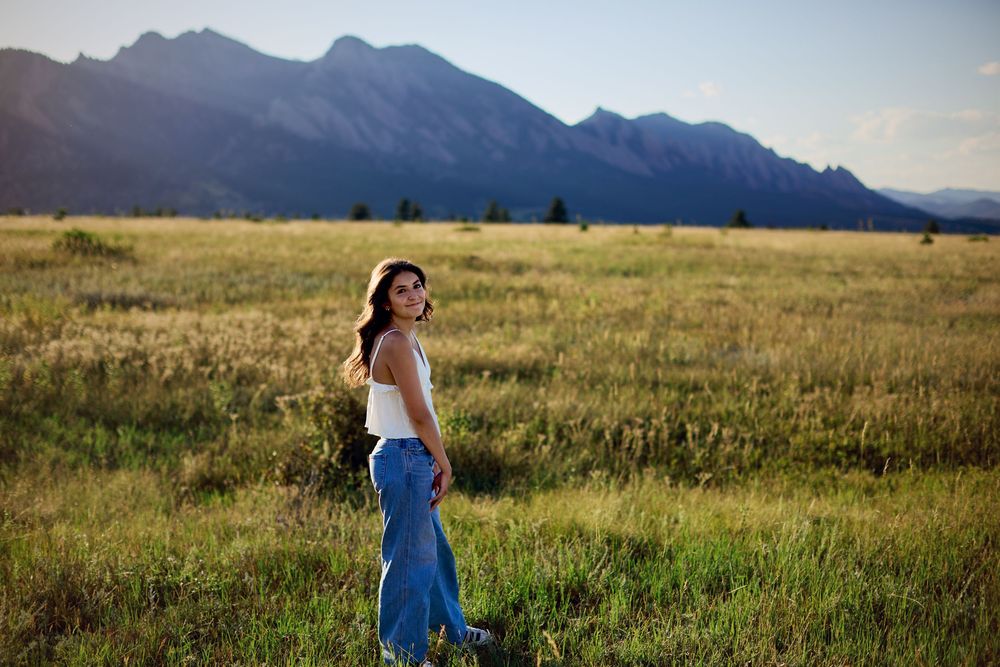 Young woman in a white top and jeans stands in a golden grassy meadow with rugged mountains and a soft sunset sky behind her.