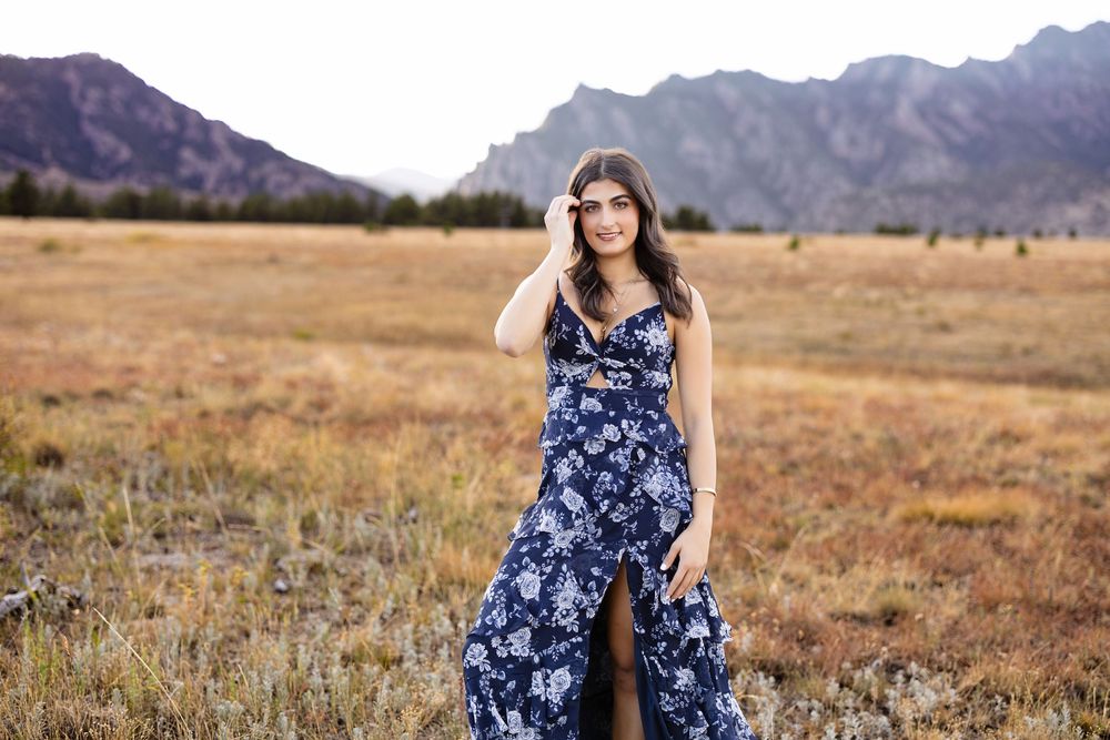 Young woman in a navy floral maxi dress with a leg slit poses in a golden grass field with rugged mountains in the background.