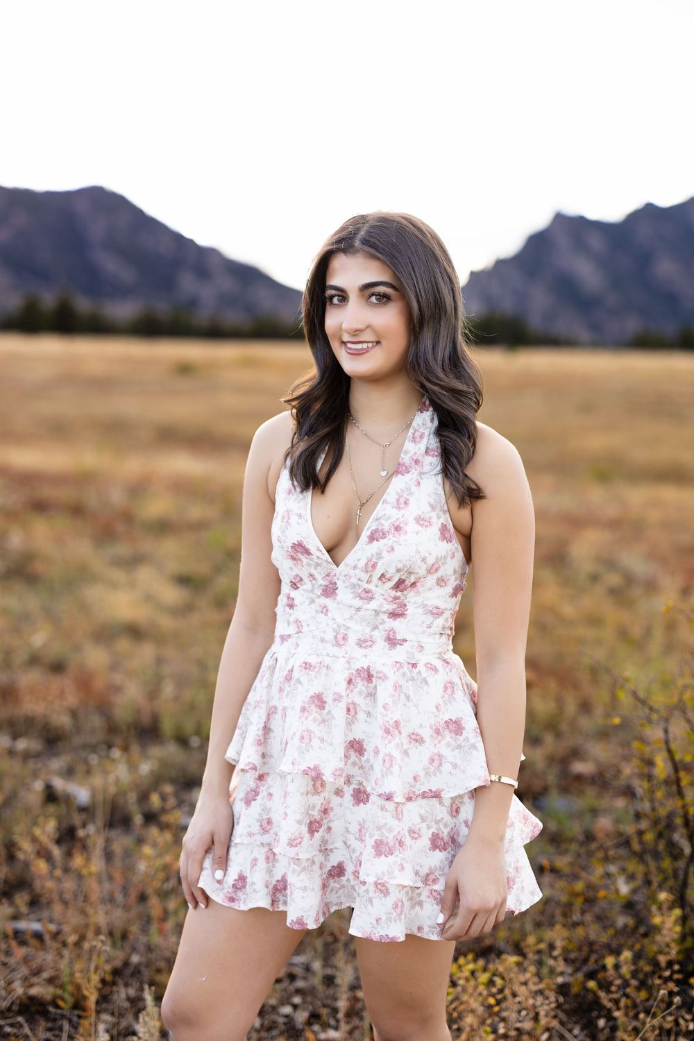 Young woman with long dark hair smiling in a pink floral ruffled sundress, standing in a golden meadow with mountains behind her.