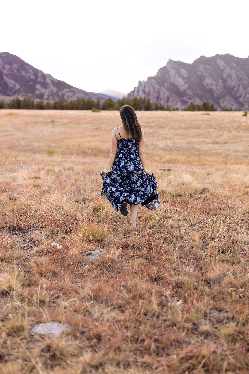Woman in a flowing navy floral sundress walks barefoot through a golden grass meadow toward rugged mountains at dusk.