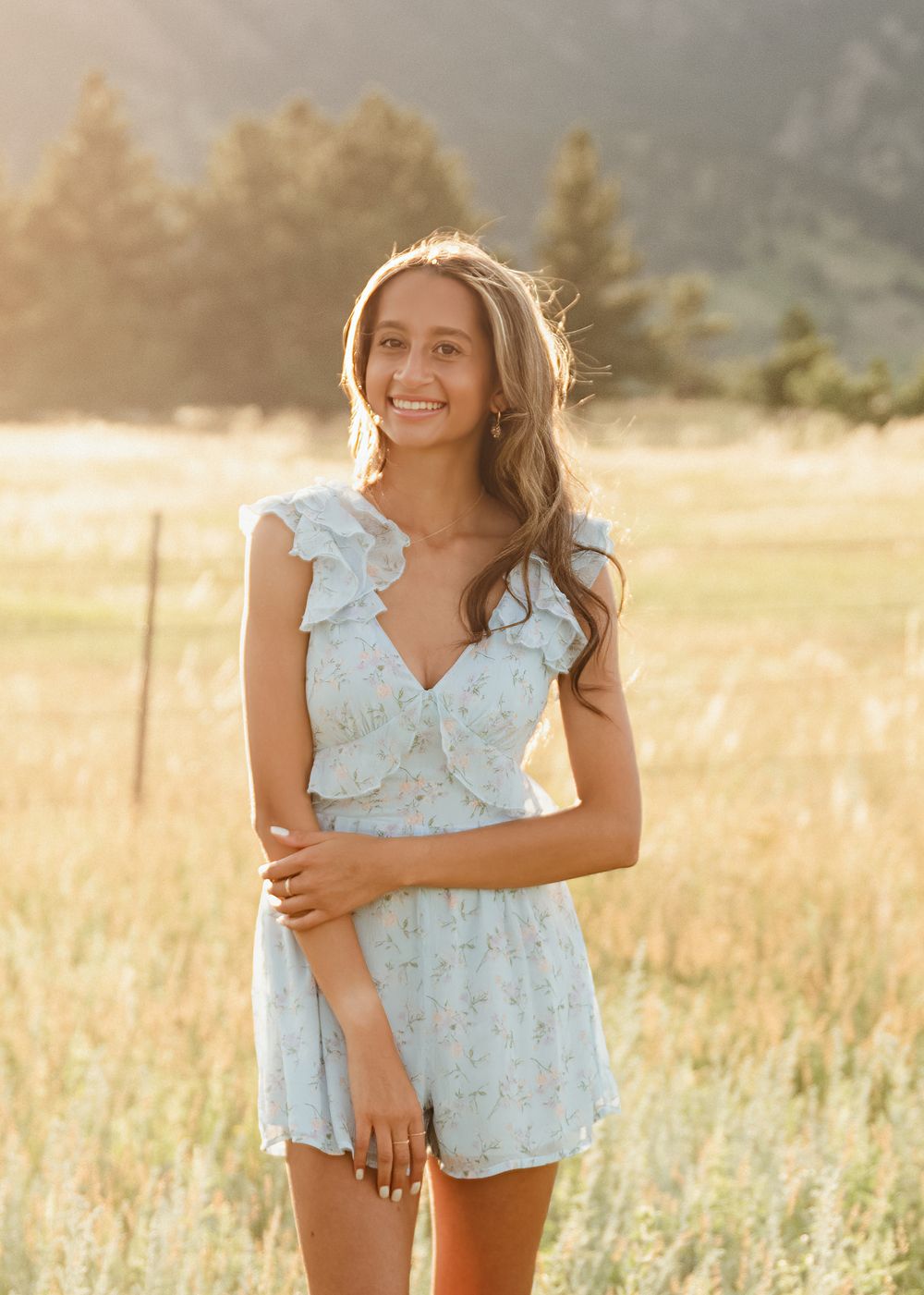 Smiling young woman in a light blue floral ruffled romper standing in a golden grassy field with trees in the soft sunset light.