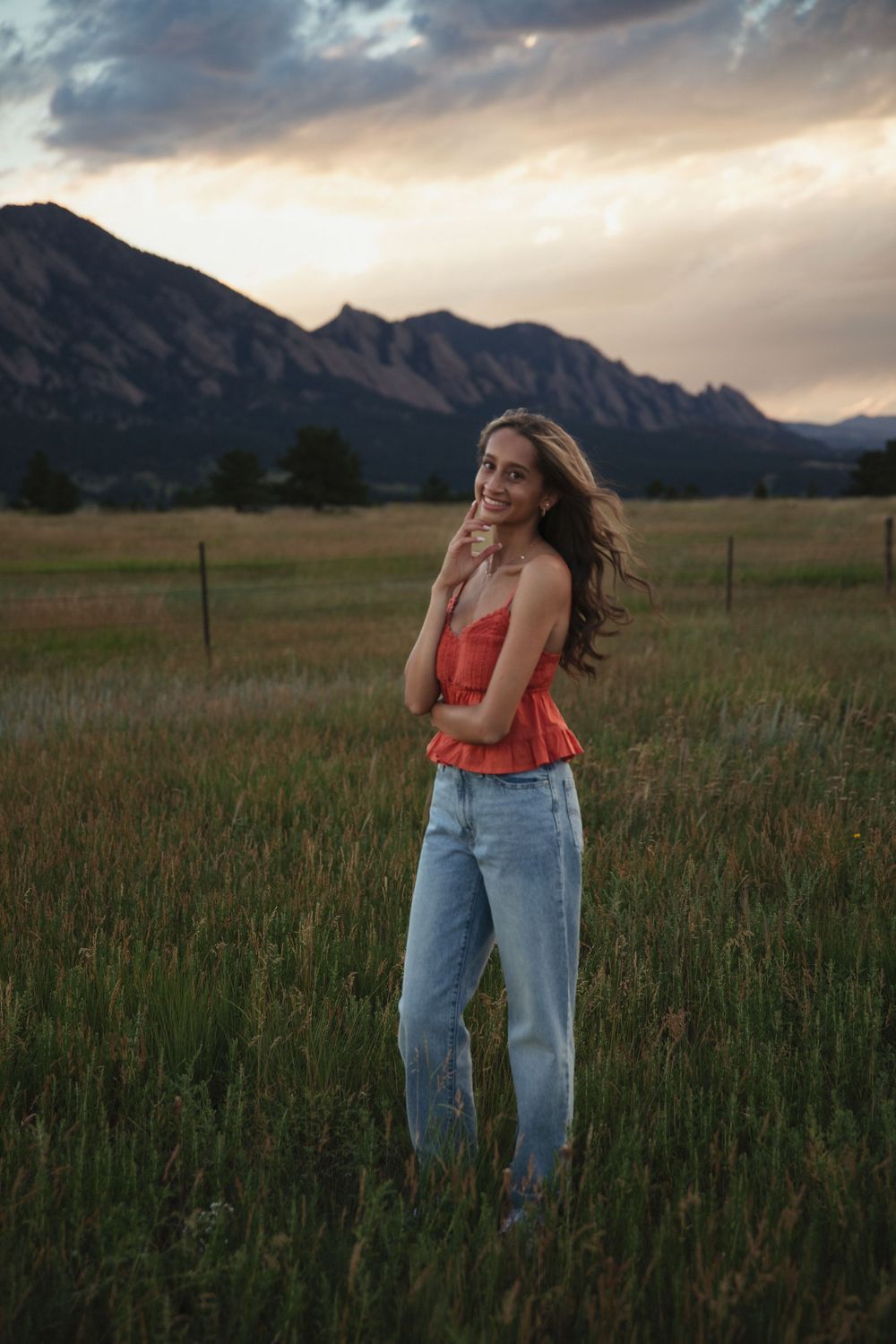 Young woman in a coral top and light-wash jeans poses in a grassy field with rugged mountains and a dramatic cloudy sunset sky behind her.
