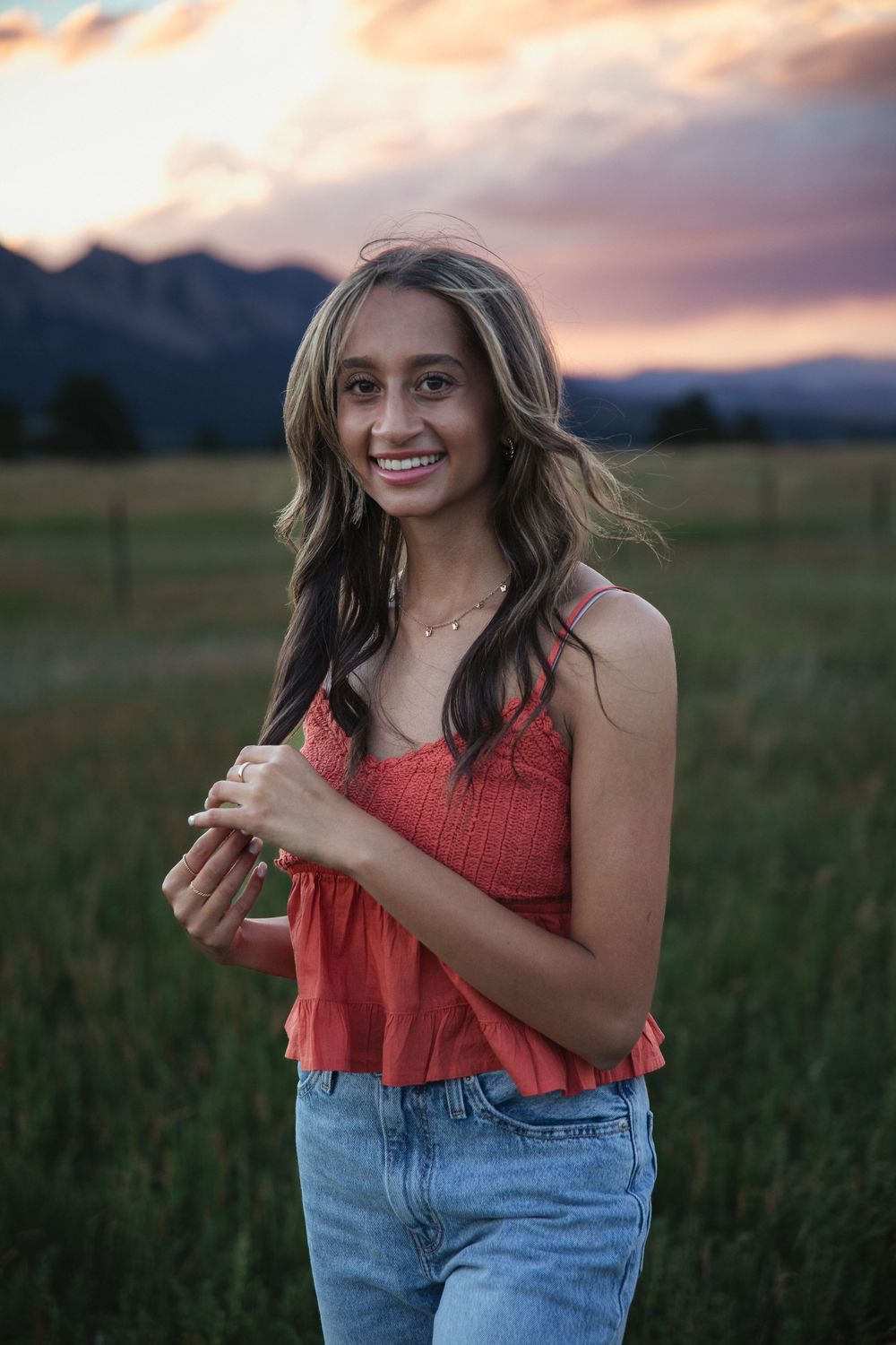 Smiling young woman in a coral top and denim jeans stands in a grassy field with mountains and a pink sunset sky behind her.
