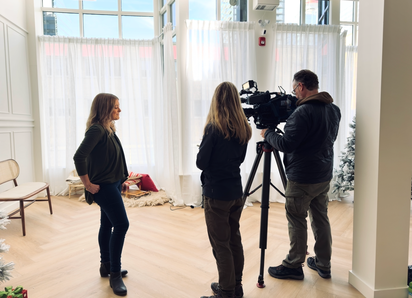 A woman stands in a bright room with large windows while a camera operator films a second woman during an on-location video interview.