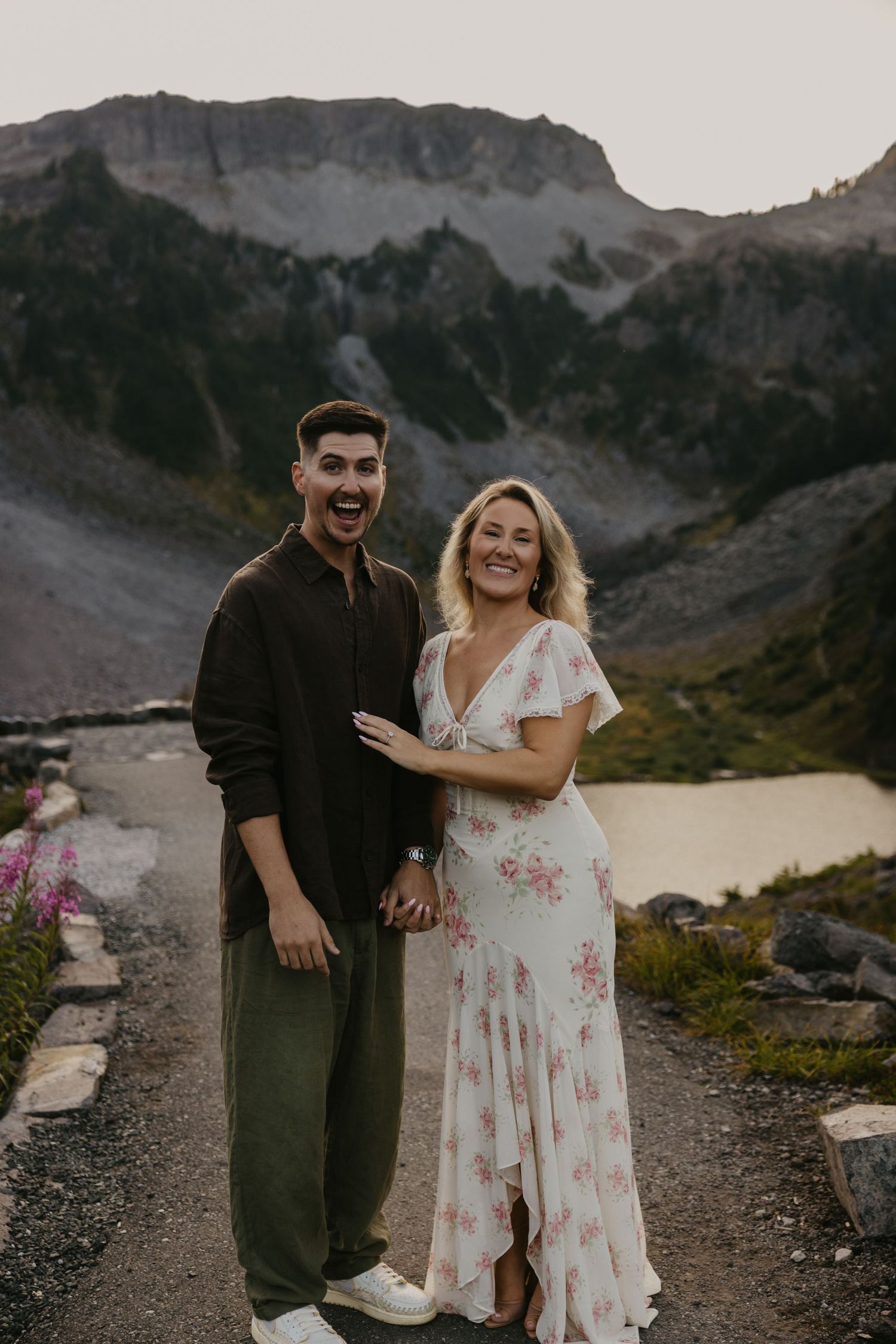 Smiling couple in a floral maxi dress and olive pants stand on a mountain trail with rugged peaks and an alpine lake behind them.