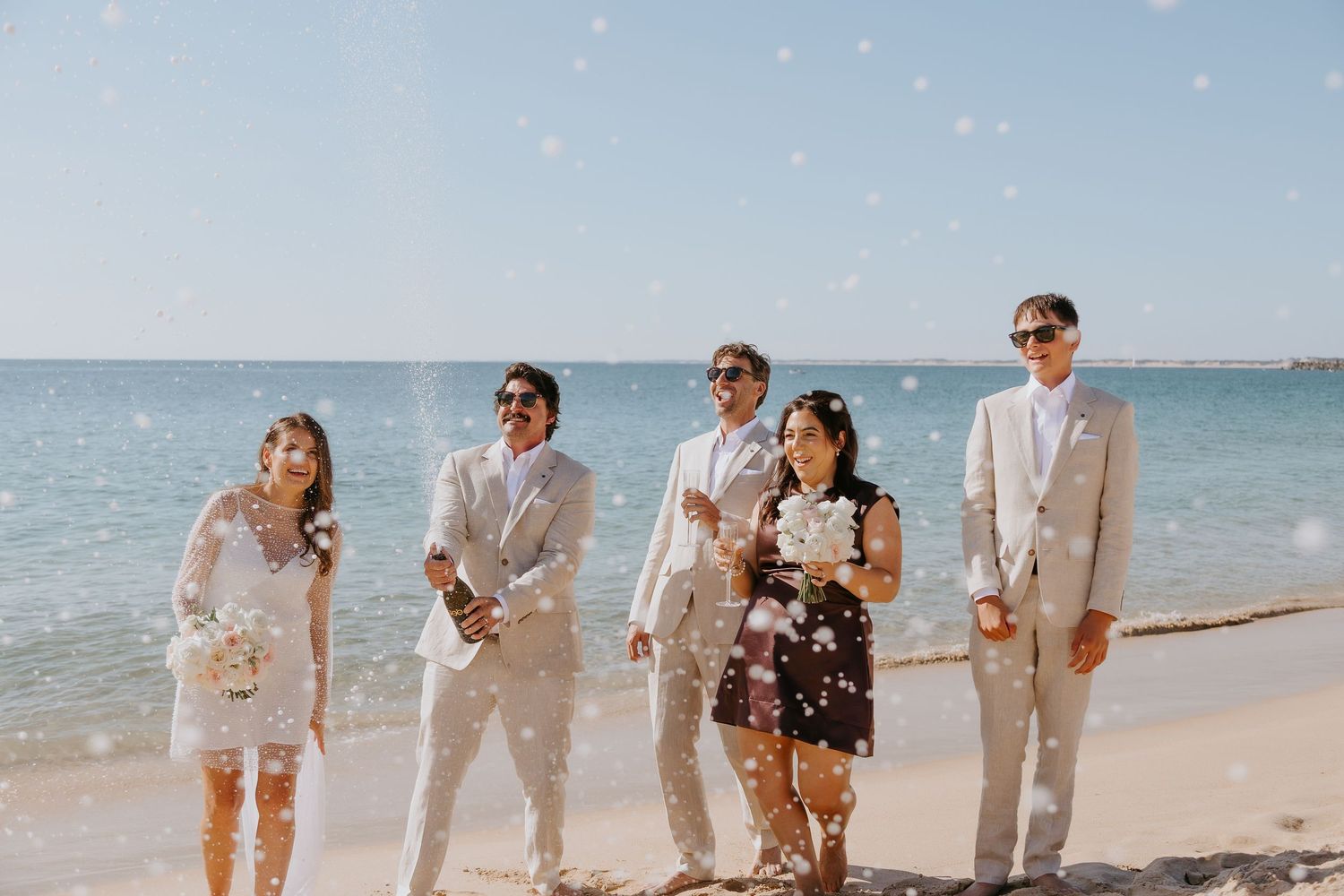Bride, groom, and wedding guests in light suits laugh and celebrate with champagne on a sunny beach while confetti falls by the ocean.