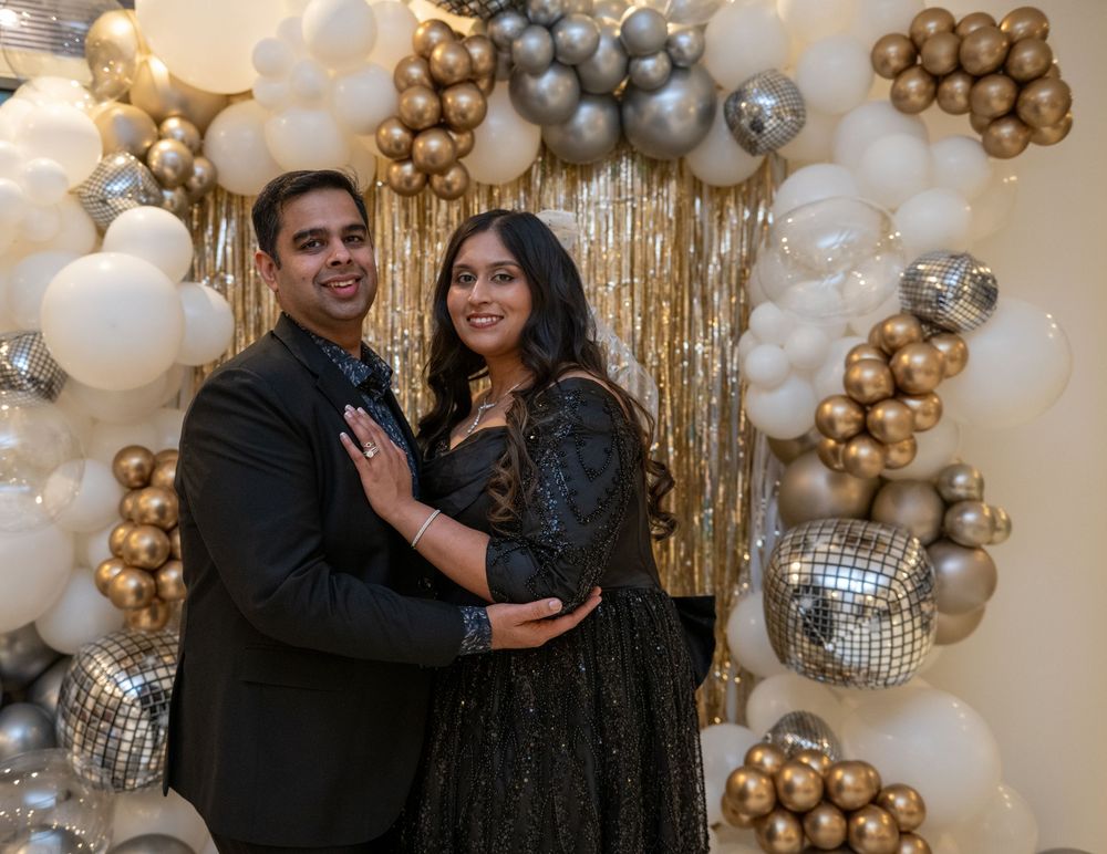Couple posing together in matching black outfits in front of elegant gold and white balloon arch.