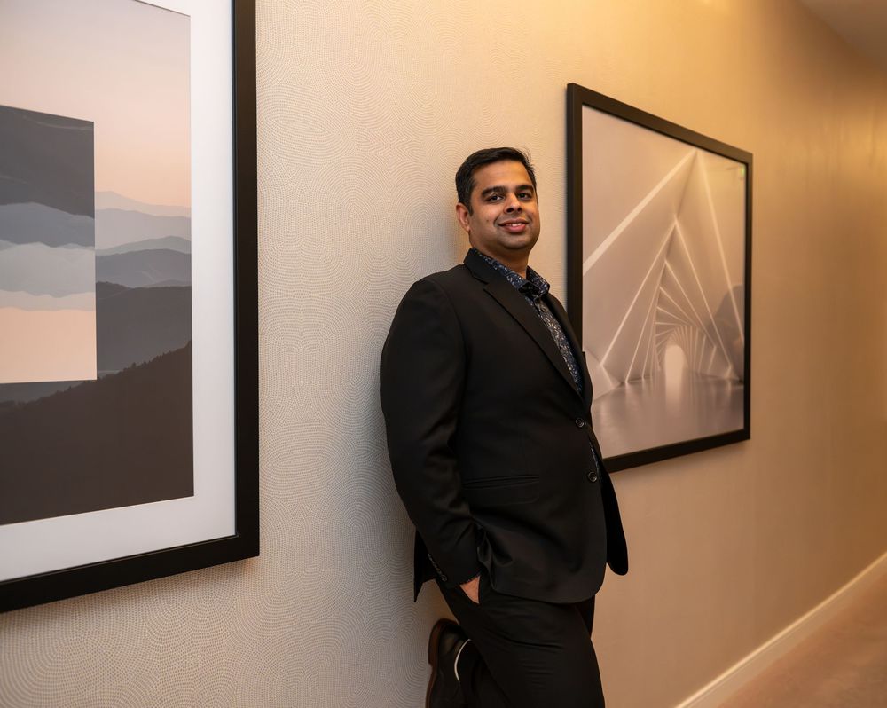 Man in black suit smiling while leaning against wall with framed artwork in elegant gallery setting.